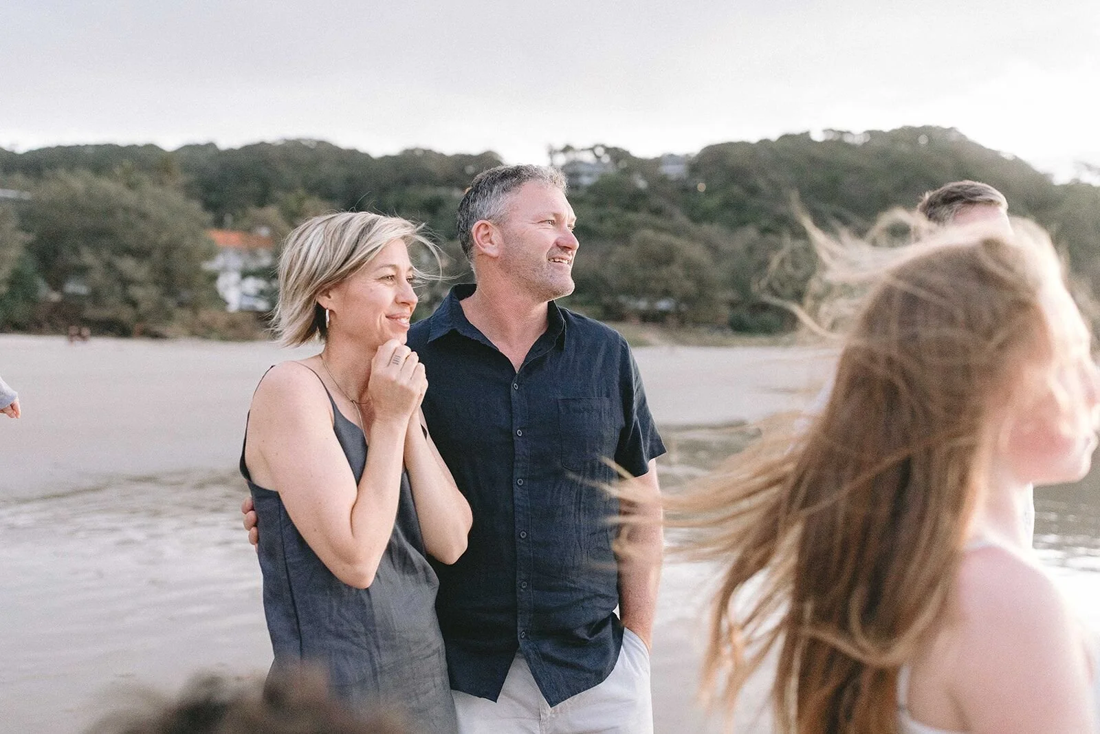 Mum and dad standing arm in arm on the beach as the wind blows their daughter's hair