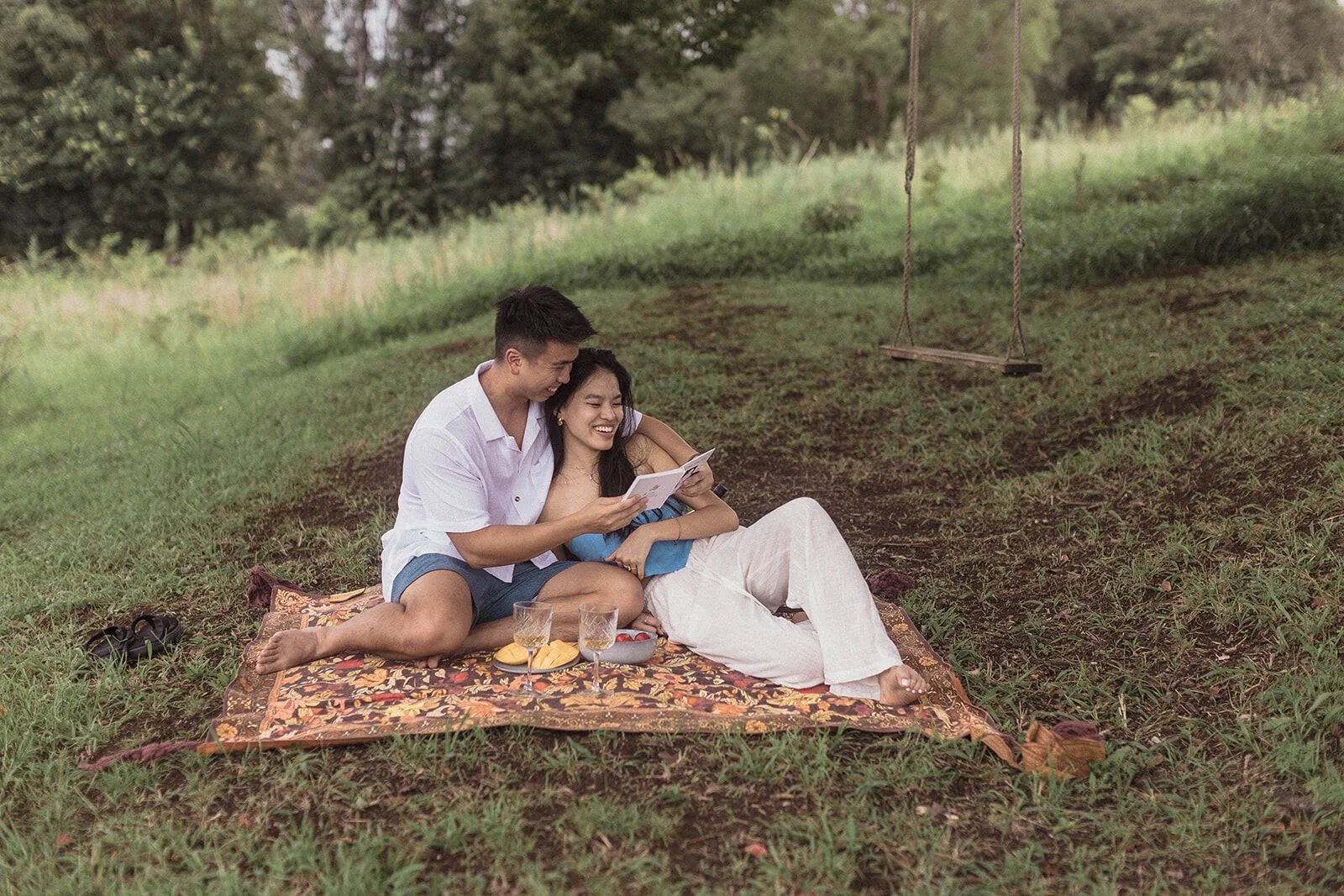 Couple snuggled together on the picnic rug reading a small book, Darien and Jacqui's picnic marriage proposal at Heartwood Farm, Byron Bay hinterland