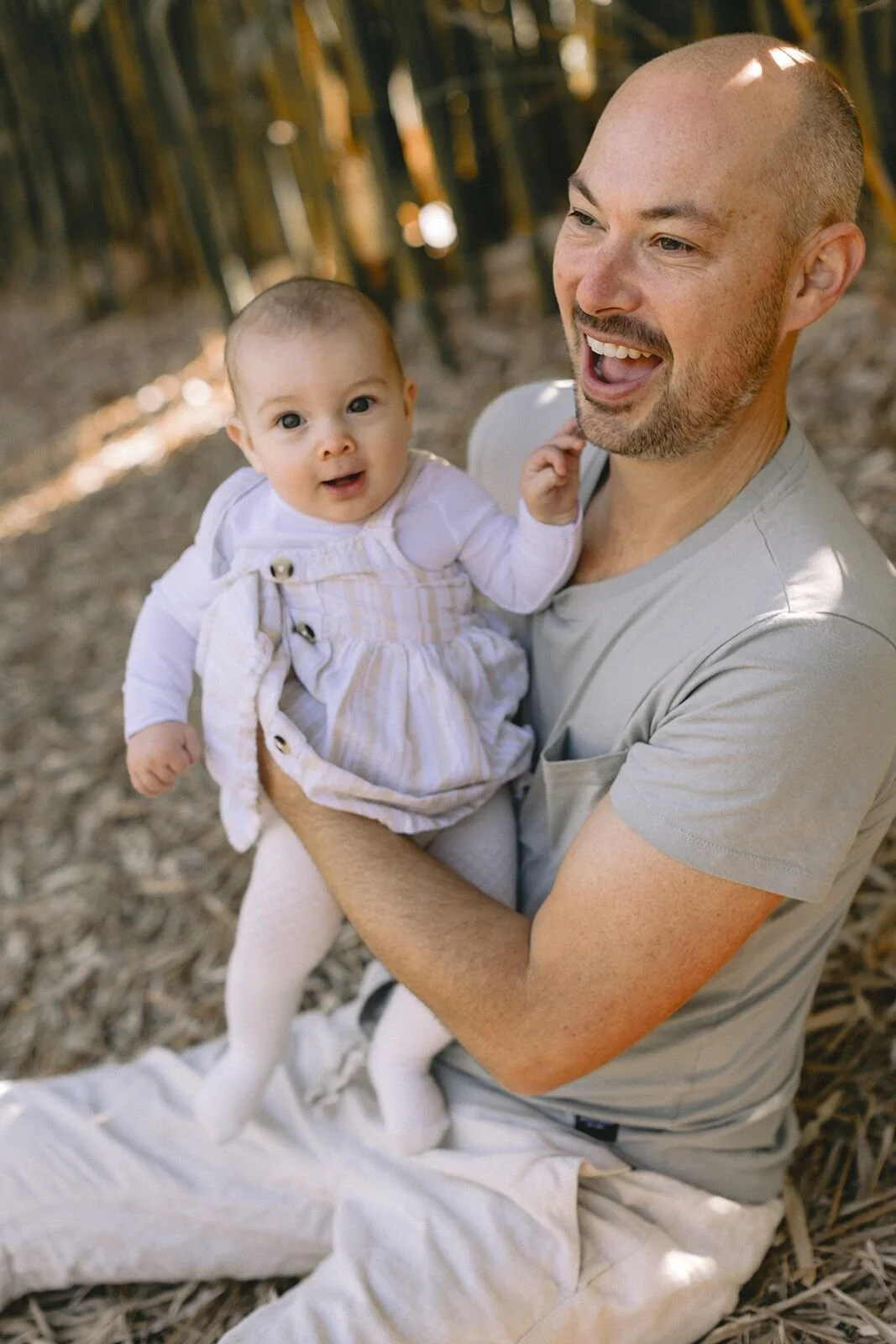 Dad laughing with his baby daughter held up on his knee, sitting among bamboo leaves