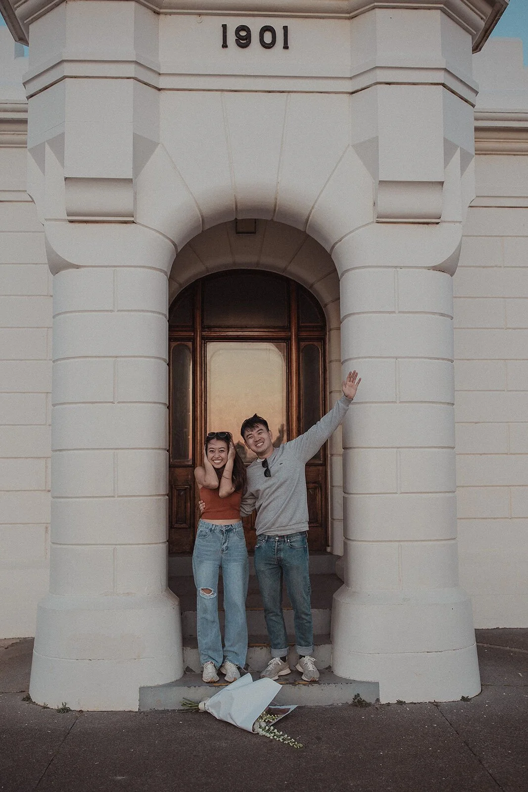 Couple celebrating in the lighthouse doorway with arms raised and the bouquet at their feet, Douglas and Ying's sunset marriage proposal at Cape Byron Lighthouse, Byron Bay