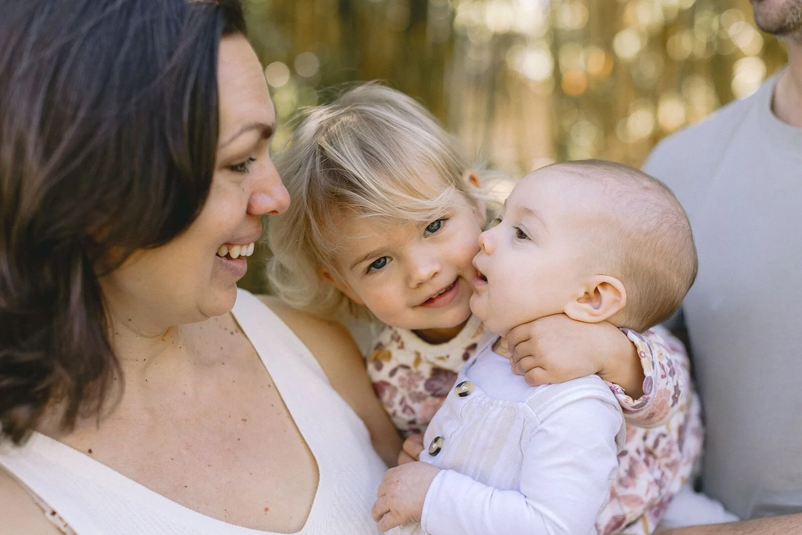 Toddler sister leaning in to kiss her baby sister while mum laughs beside them