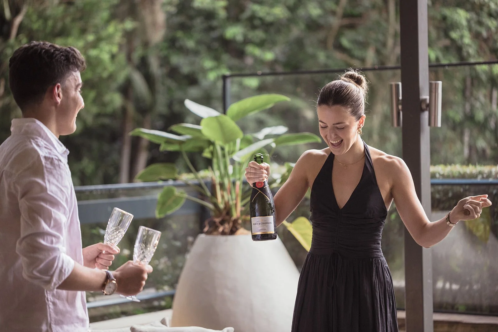 Woman laughing and holding the champagne bottle as her fiancé holds the glasses, Ben and Chloe's marriage proposal in Byron Bay