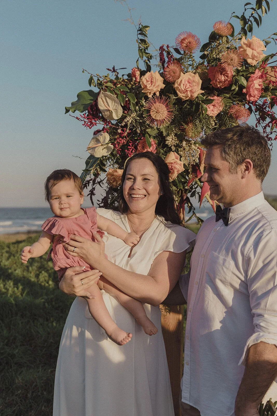 Bride holding a baby beside the groom in front of the flowers, Maritess and Mark's elopement at Lennox Head