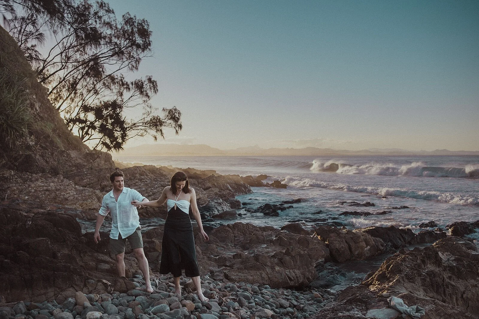 Man helping his fiancée across the rocks, John and Emma's sunset marriage proposal at Wategos Beach, Byron Bay