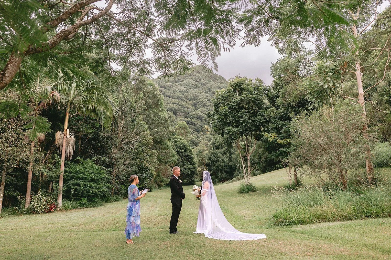 Couple and celebrant on a lawn surrounded by rainforest hills, Kobi and Darren's elopement at Butterfly Lane, Byron Bay hinterland
