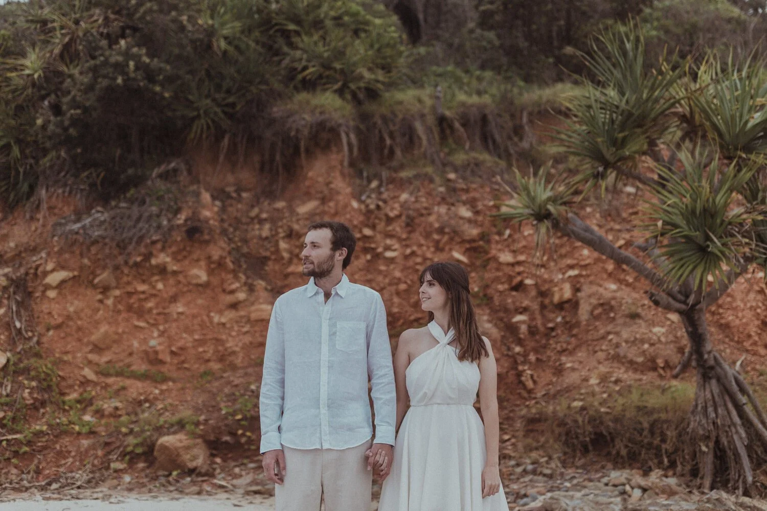 Couple standing in front of red cliffs and pandanus trees, Yoanne and Elisa's rainy elopement at Broken Head, Byron Bay