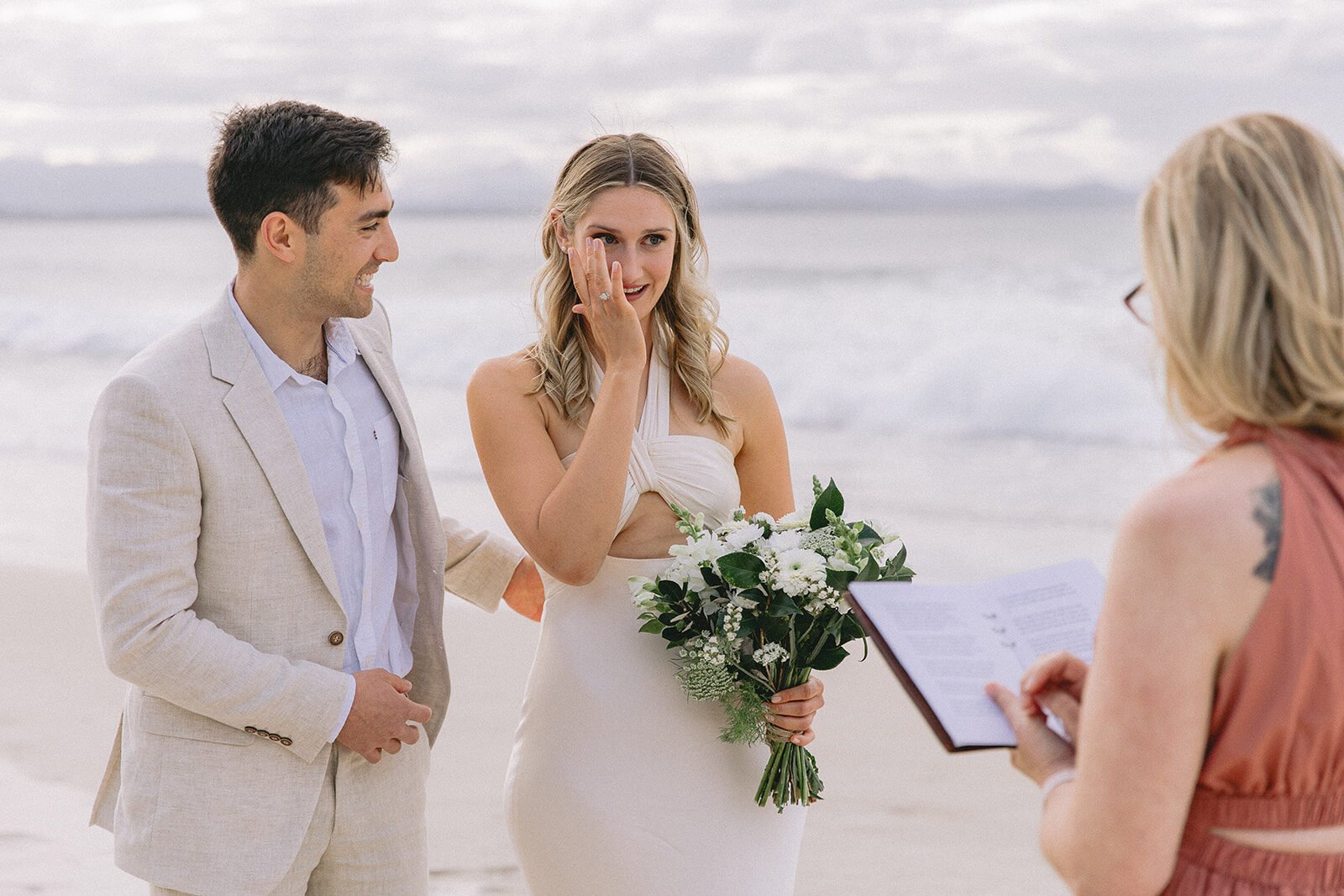Bride wiping a happy tear as the groom and celebrant look on, Abby and James' elopement at Little Wategos Beach, Byron Bay