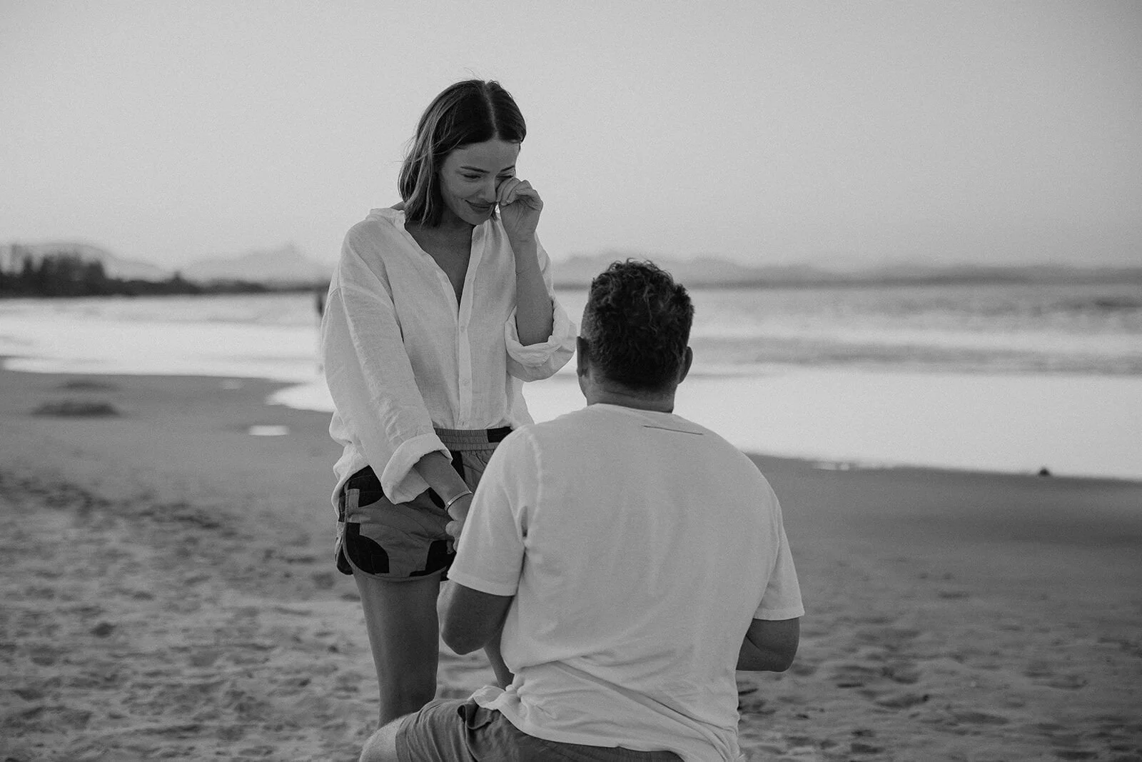 Black and white of the woman wiping a tear as her partner kneels, Jamie and Jessica's beach marriage proposal at Belongil Beach, Byron Bay