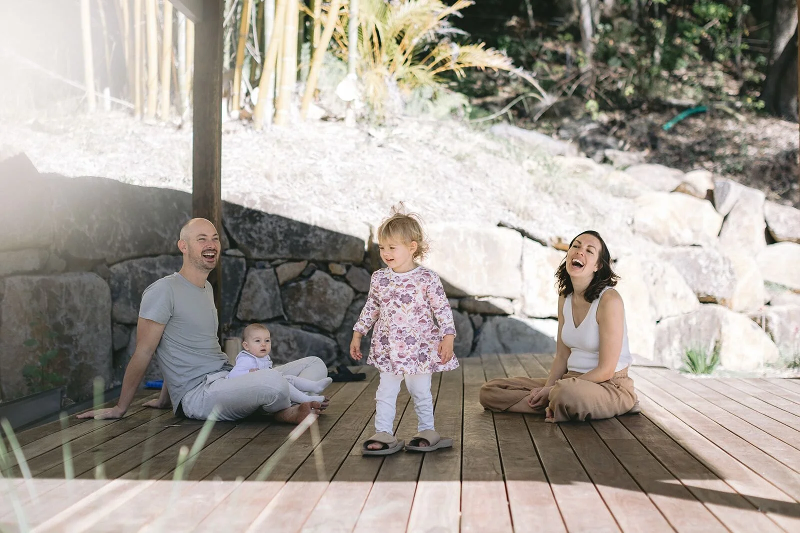 Family relaxing on a timber deck, toddler standing between mum and dad, baby on dad's lap