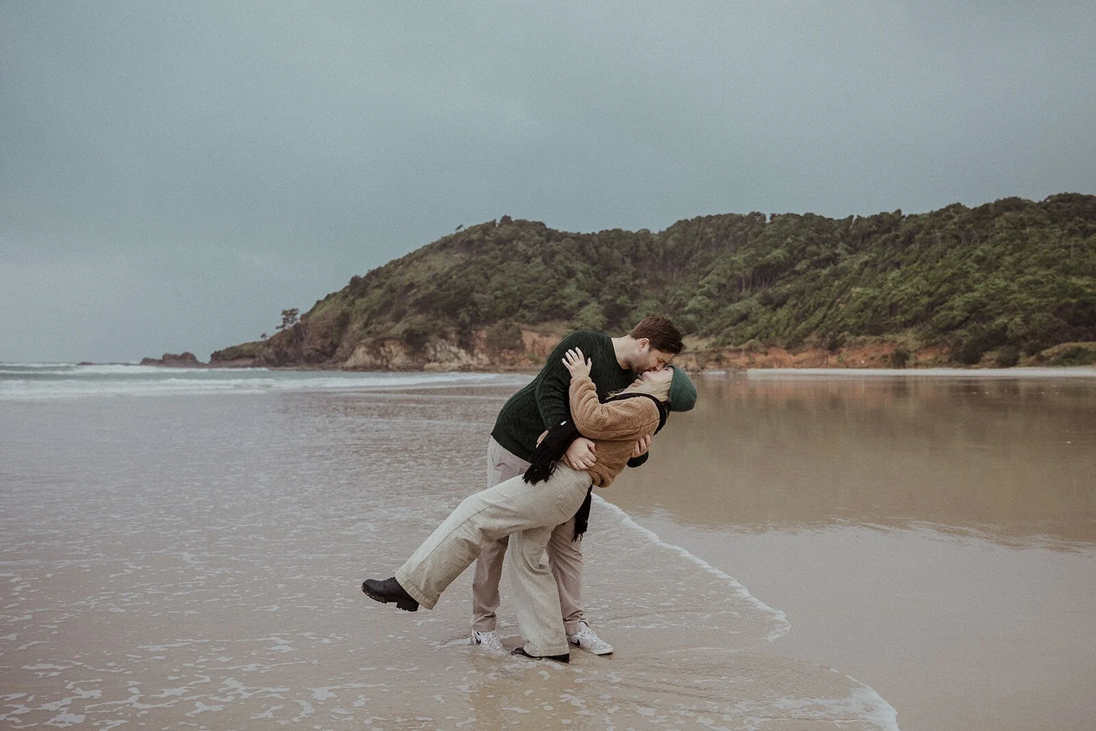 Couple dipping into a kiss at the shoreline with the headland behind, Raphael and Vany's rainy day marriage proposal at Broken Head Beach, Byron Bay