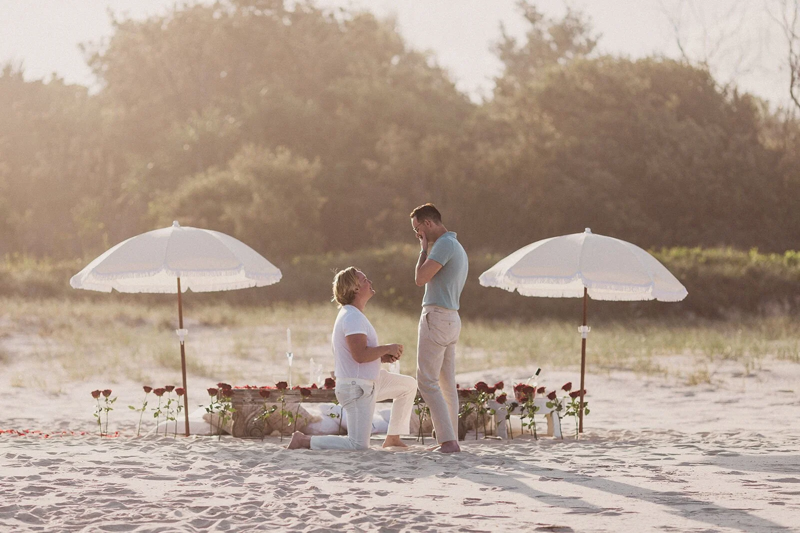 Man kneeling on the sand to propose beside the rose-lined beach picnic, Byron and Luke's marriage proposal at Cabarita Beach, Northern Rivers