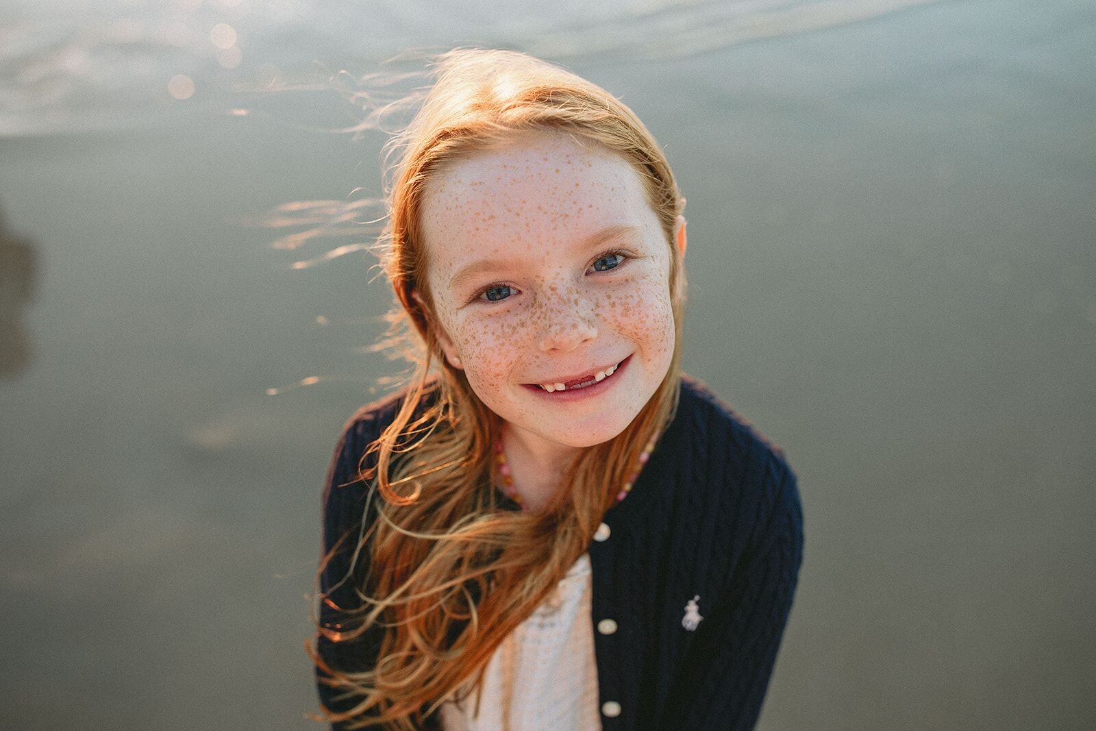 Close-up portrait of a smiling girl with freckles and windblown red hair at the water's edge
