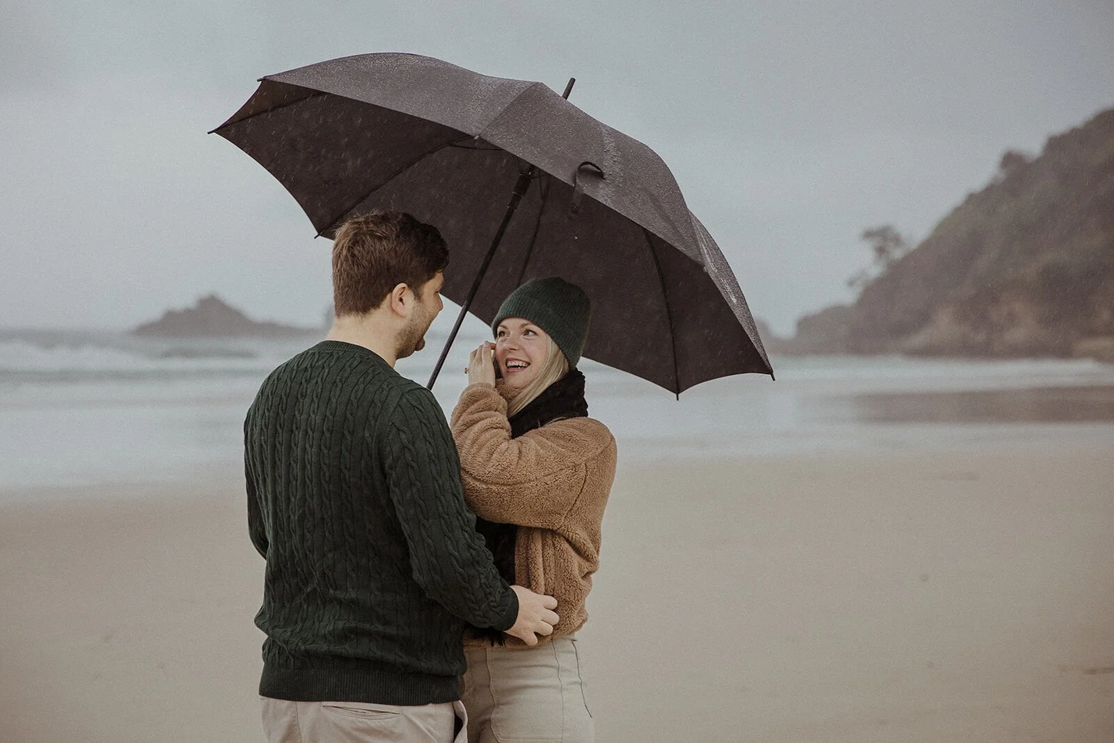 Woman laughing and wiping a tear as her fiancé holds her under the umbrella, Raphael and Vany's rainy day marriage proposal at Broken Head Beach, Byron Bay