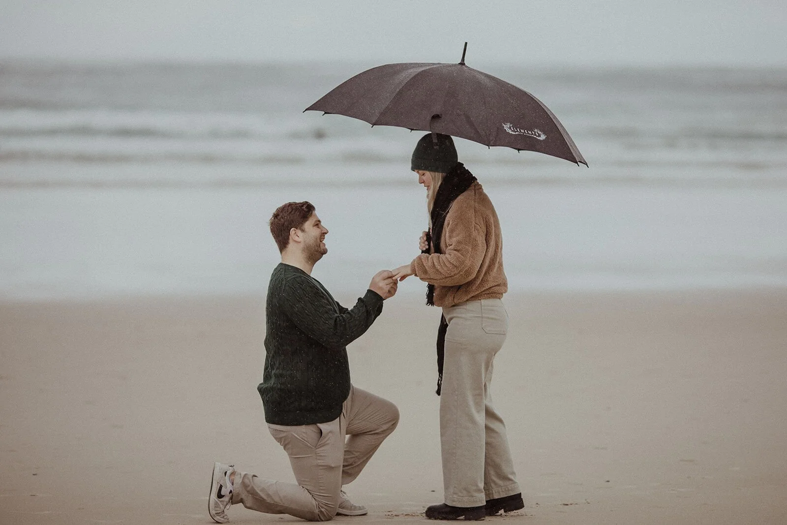 Man kneeling on the wet sand holding his partner's hand to propose beneath an umbrella, Raphael and Vany's rainy day marriage proposal at Broken Head Beach, Byron Bay