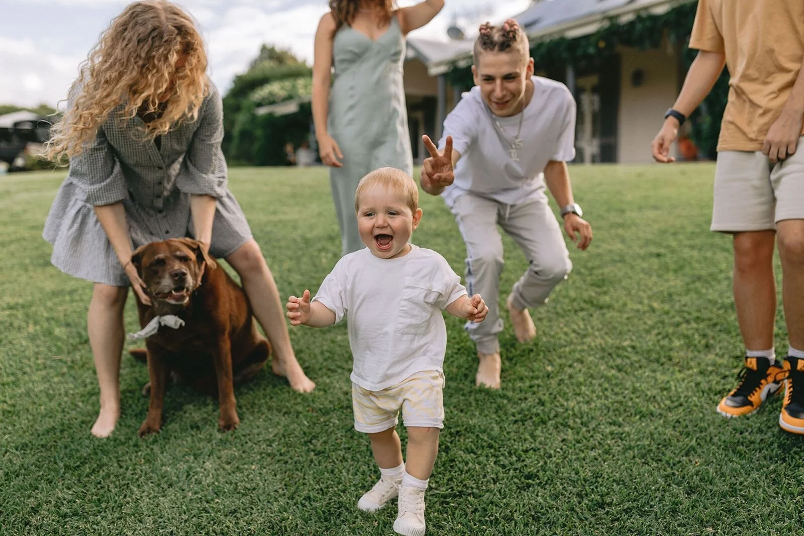 Toddler running across the lawn laughing as the family and their brown dog play behind him