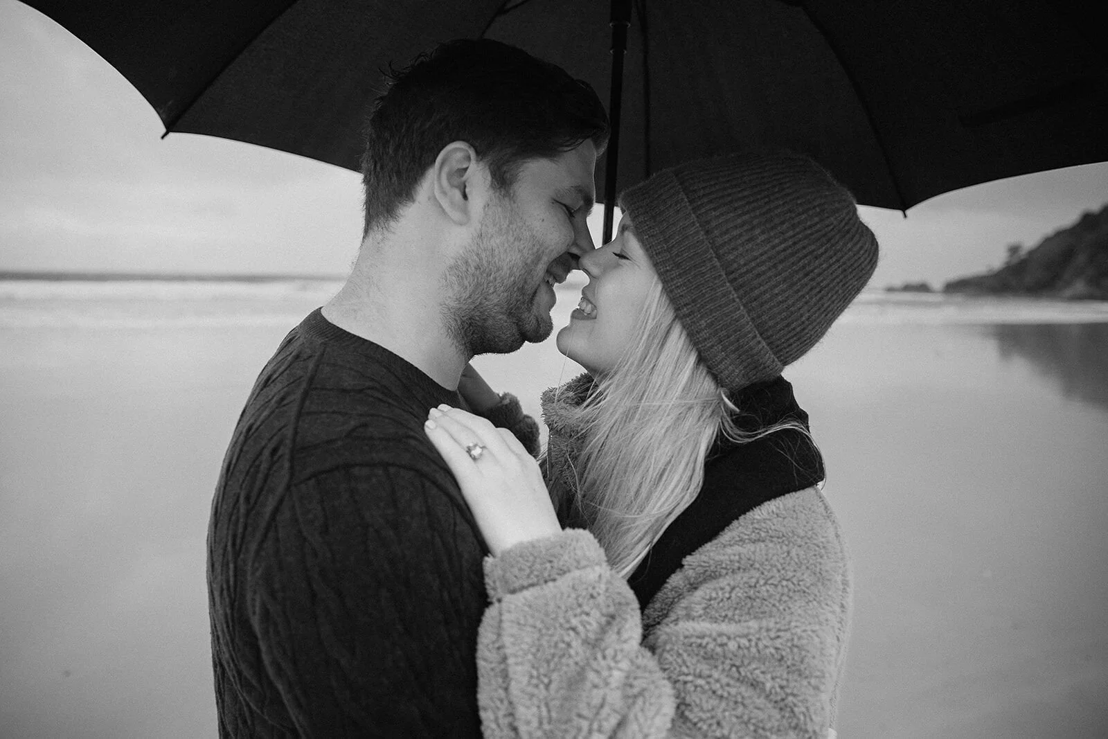 Black and white of the couple nose to nose under the umbrella, Raphael and Vany's rainy day marriage proposal at Broken Head Beach, Byron Bay