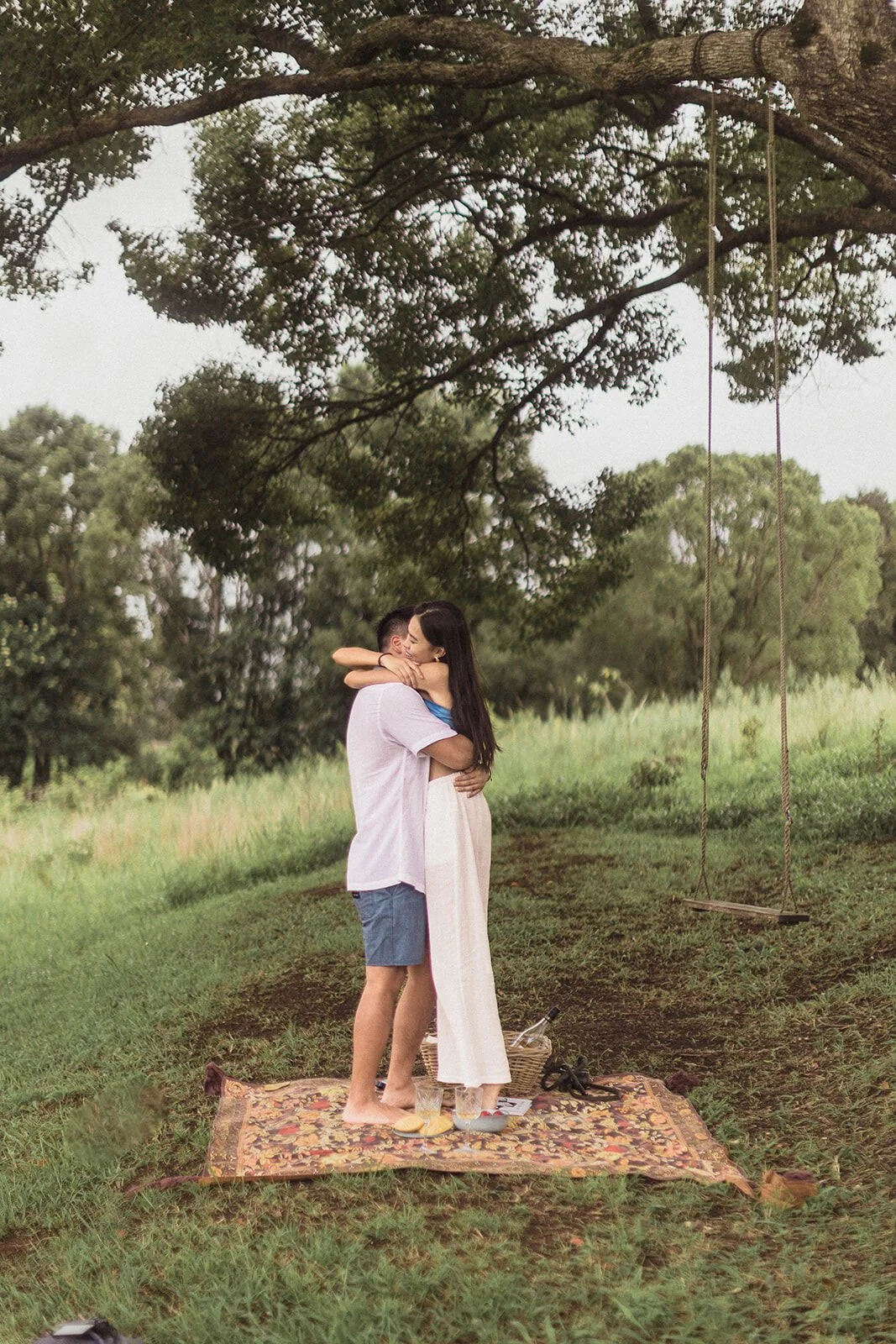 Couple hugging on the picnic rug after the proposal with the rope swing behind, Darien and Jacqui's picnic marriage proposal at Heartwood Farm, Byron Bay hinterland