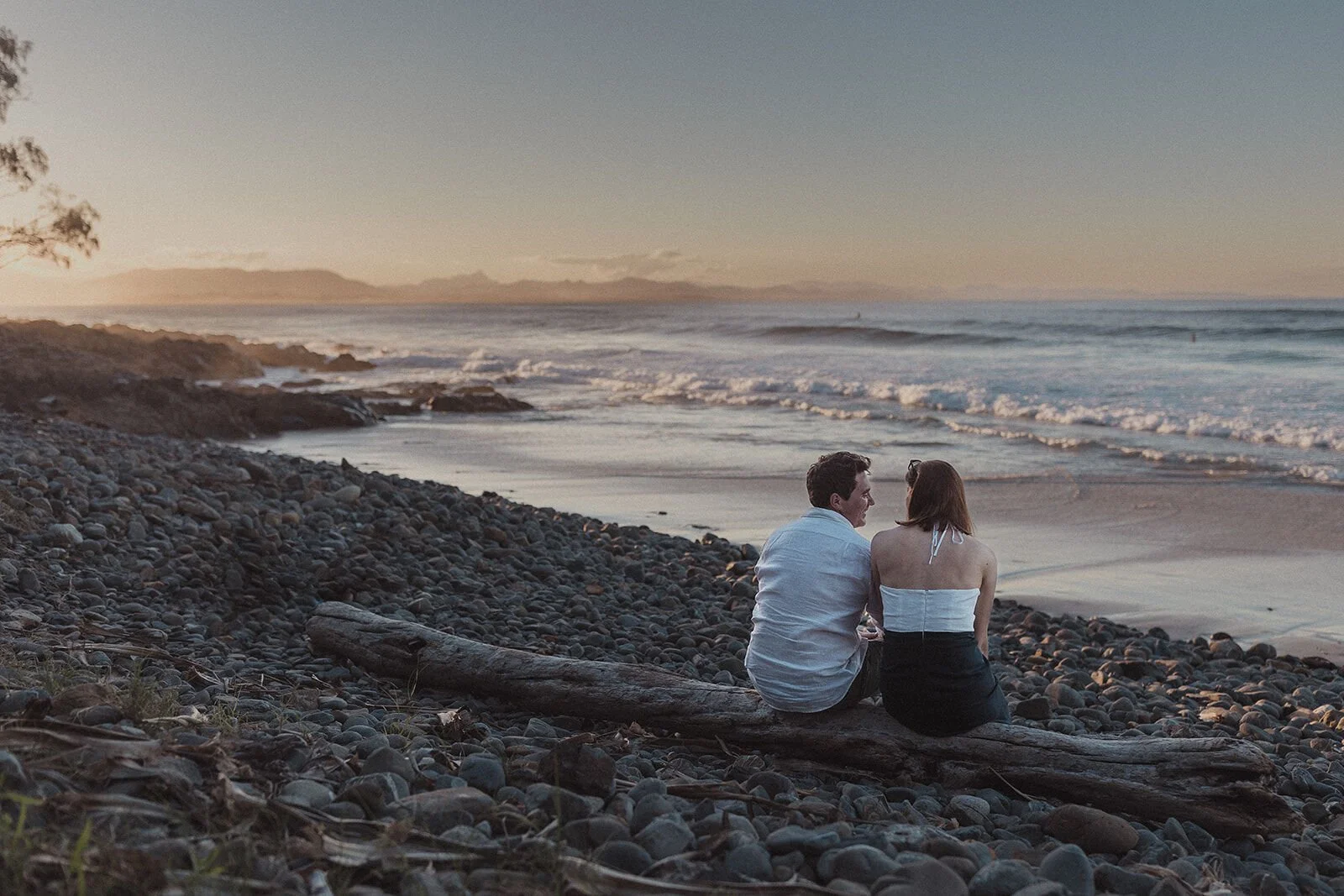 Couple sitting on a driftwood log watching the waves in the sunset, John and Emma's sunset marriage proposal at Wategos Beach, Byron Bay