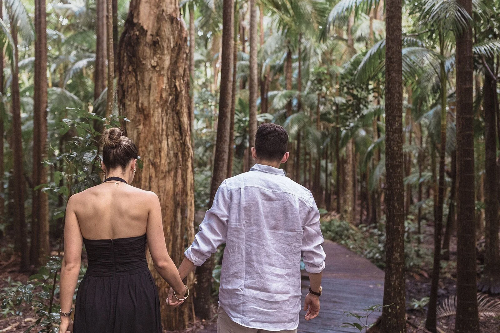 Couple from behind holding hands on the forest boardwalk, Ben and Chloe's marriage proposal in Byron Bay