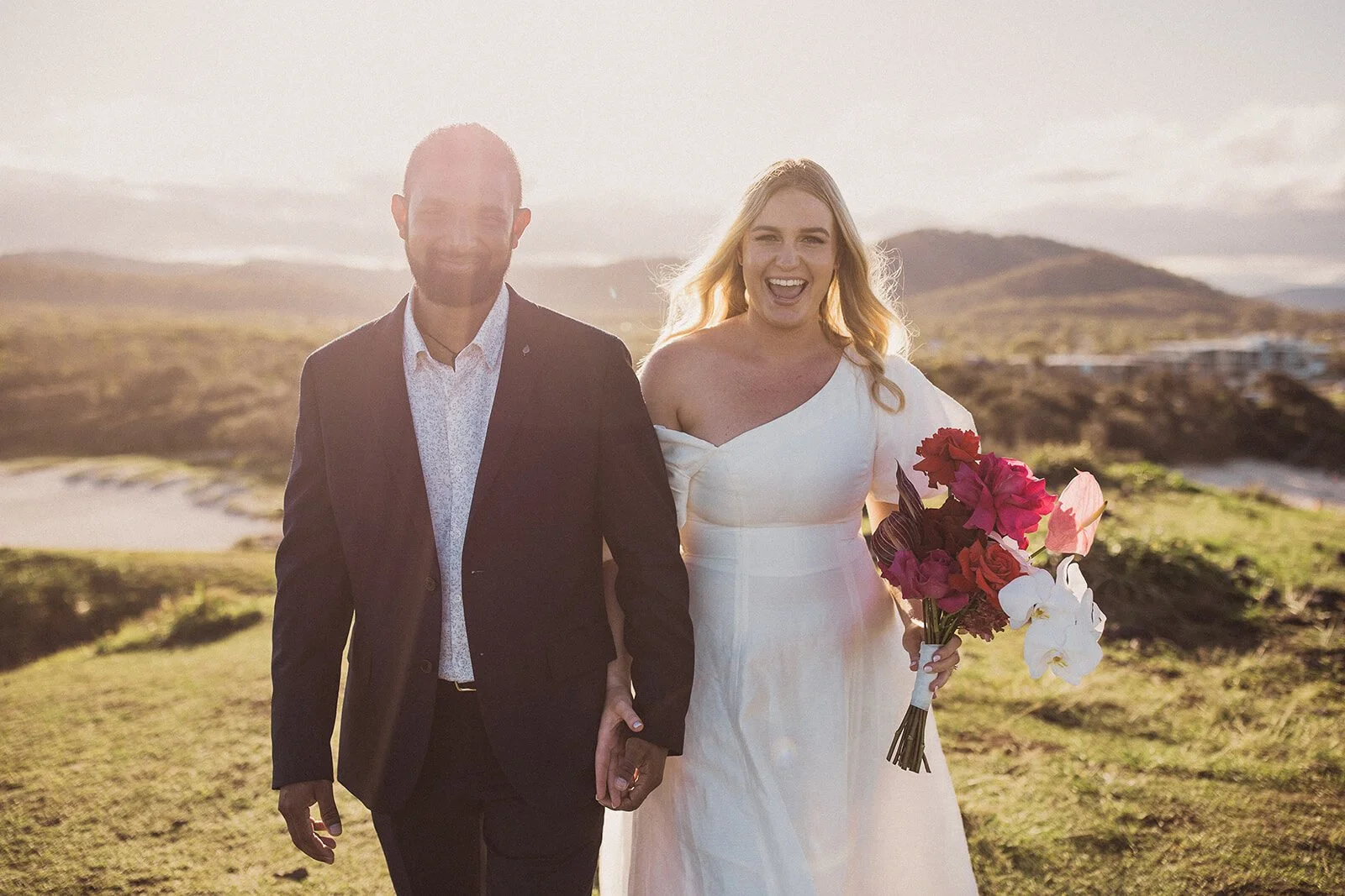 Couple laughing and walking toward the camera, bride holding a bright pink bouquet, Brittany and Mark's elopement at Cabarita Beach, Northern Rivers