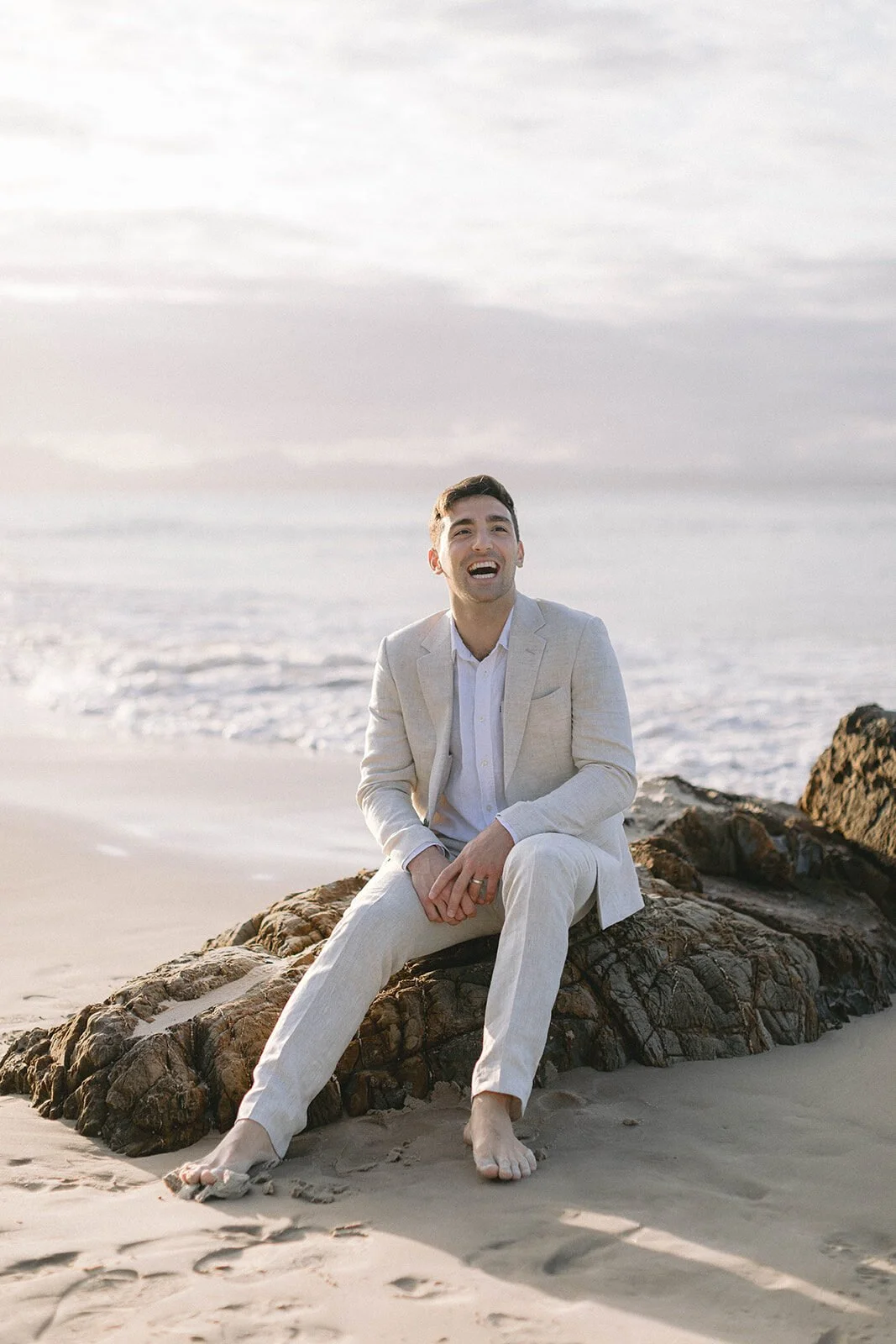 Groom laughing as he sits on a rock on the beach, Abby and James' elopement at Little Wategos Beach, Byron Bay