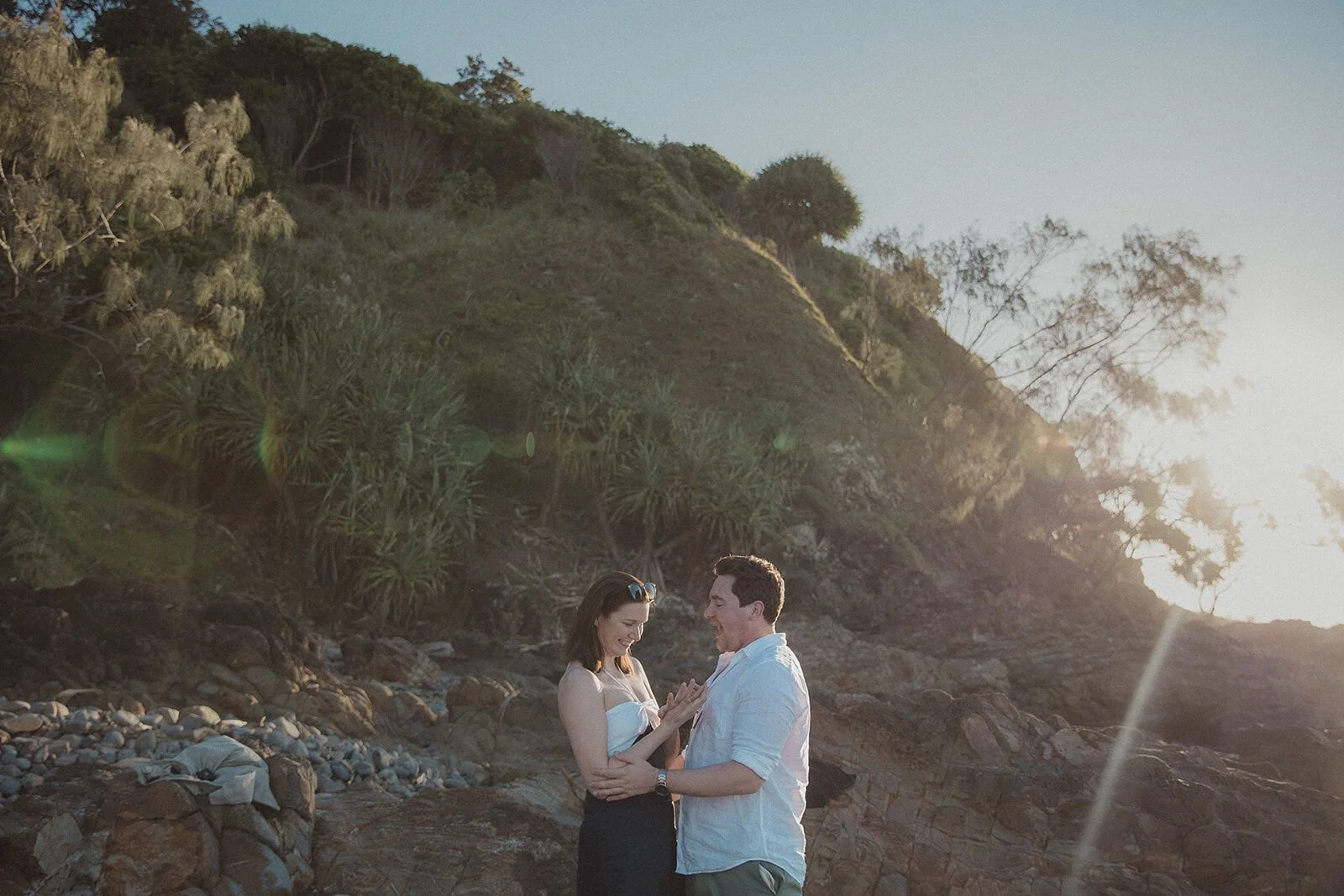 Couple embracing beneath the grassy headland with sun flare, John and Emma's sunset marriage proposal at Wategos Beach, Byron Bay