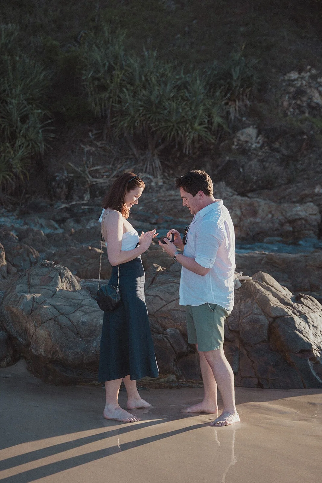 Couple looking down at the ring box on the rocks, John and Emma's sunset marriage proposal at Wategos Beach, Byron Bay