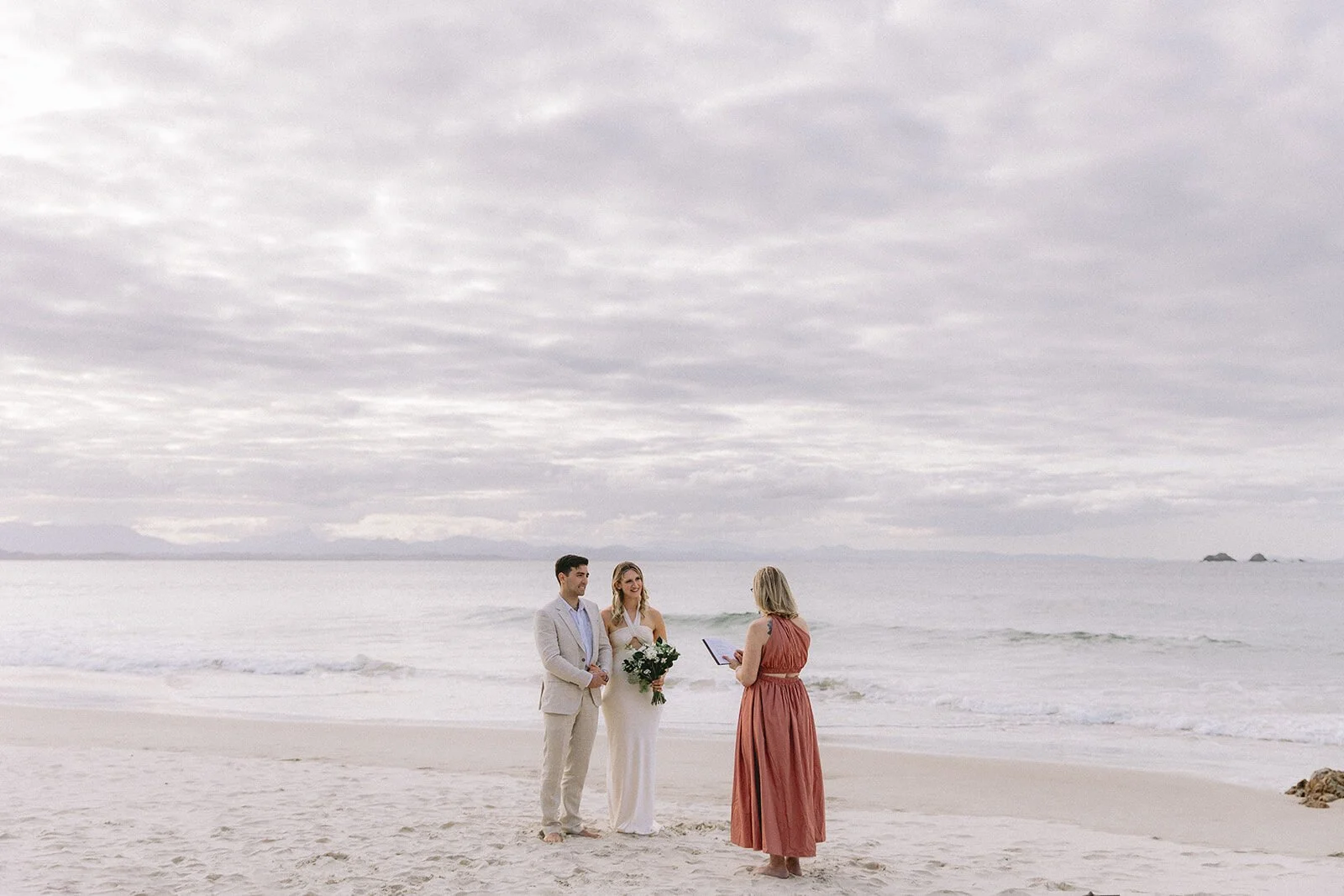 Wide view of the couple and celebrant on the empty beach, Abby and James' elopement at Little Wategos Beach, Byron Bay