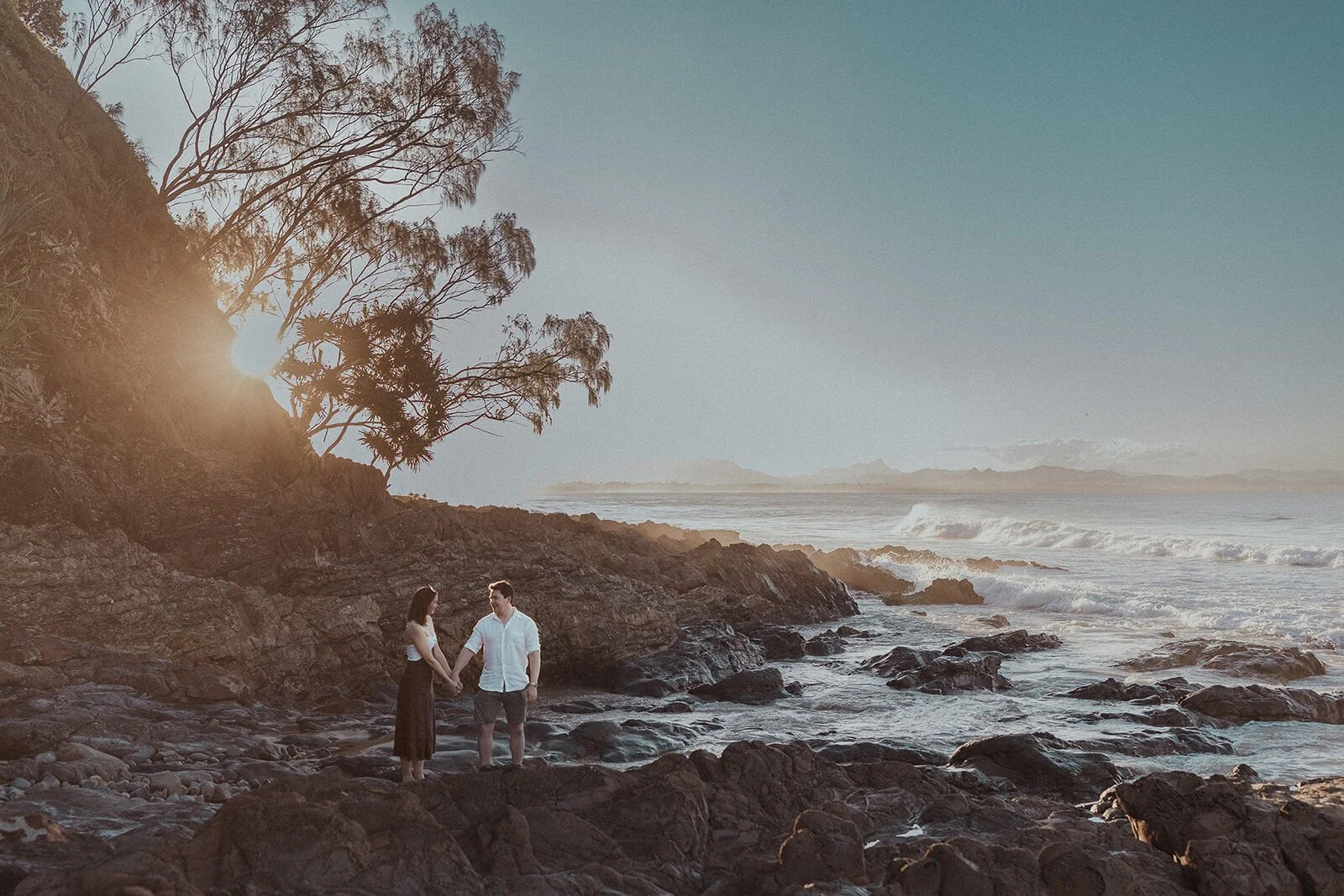 Couple holding hands on the rocks above the waves, backlit by the sunset, John and Emma's sunset marriage proposal at Wategos Beach, Byron Bay