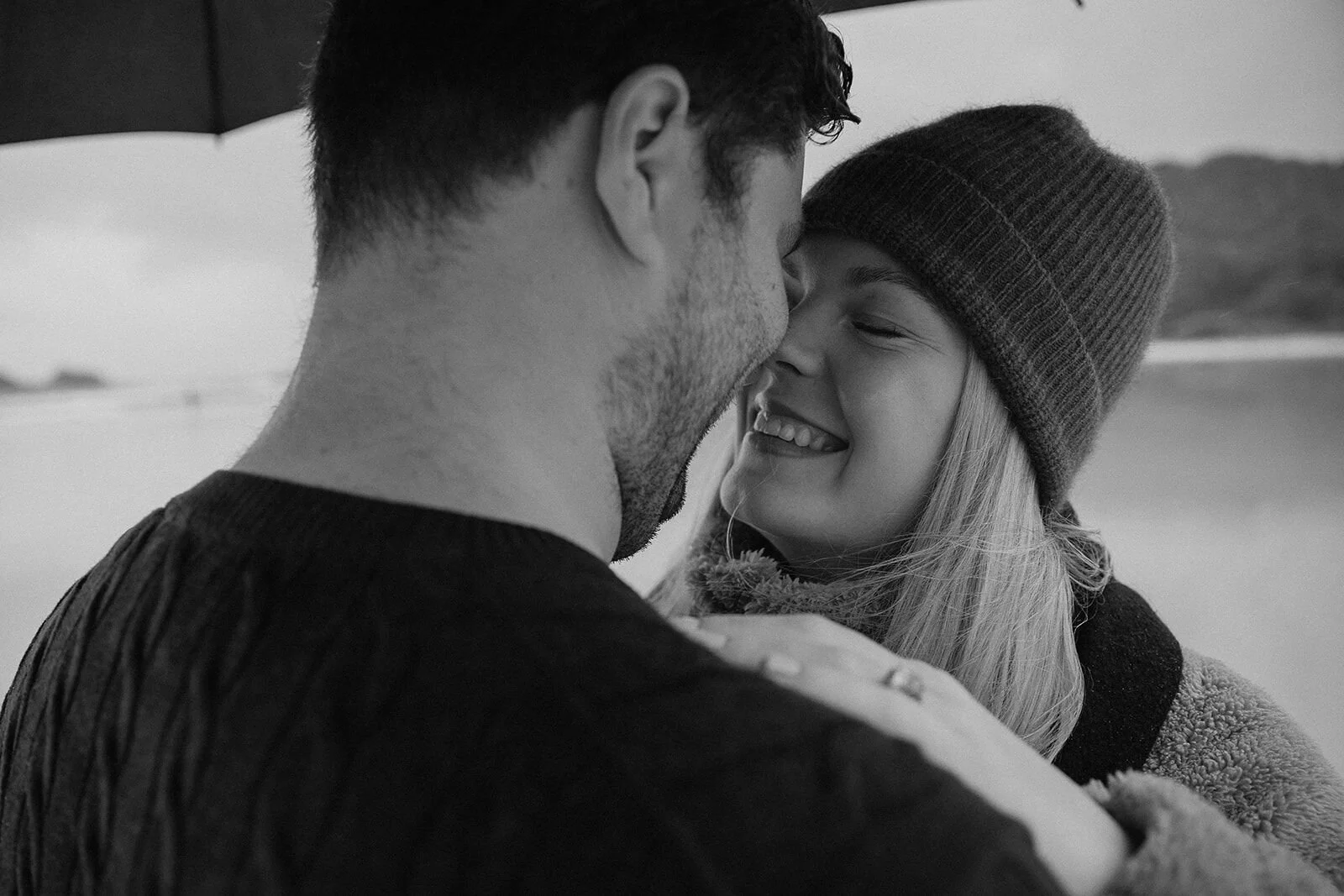 Black and white close up of the couple nose to nose and smiling, her ring on his shoulder, Raphael and Vany's rainy day marriage proposal at Broken Head Beach, Byron Bay