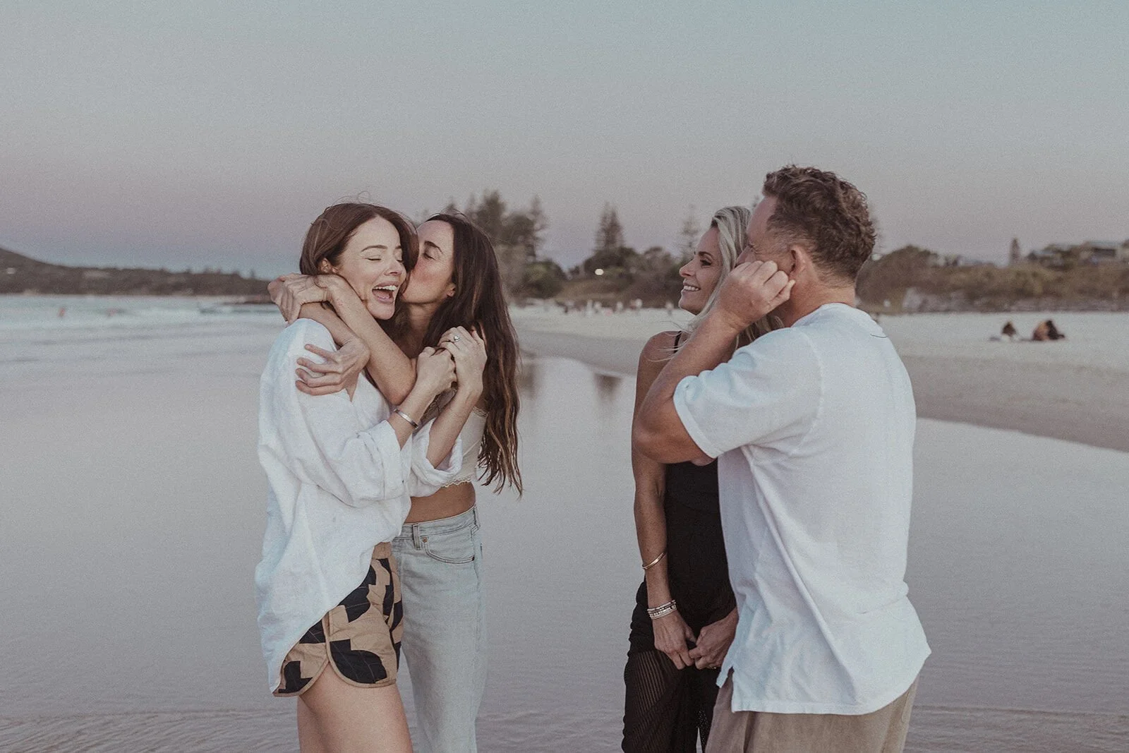 Friends kissing and hugging the bride-to-be as the couple celebrate on the shore, Jamie and Jessica's beach marriage proposal at Belongil Beach, Byron Bay