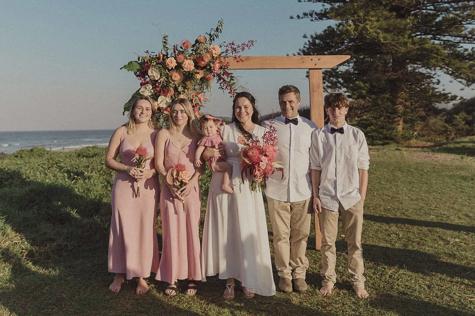 Family and wedding party posing beneath the floral arch, Maritess and Mark's elopement at Lennox Head