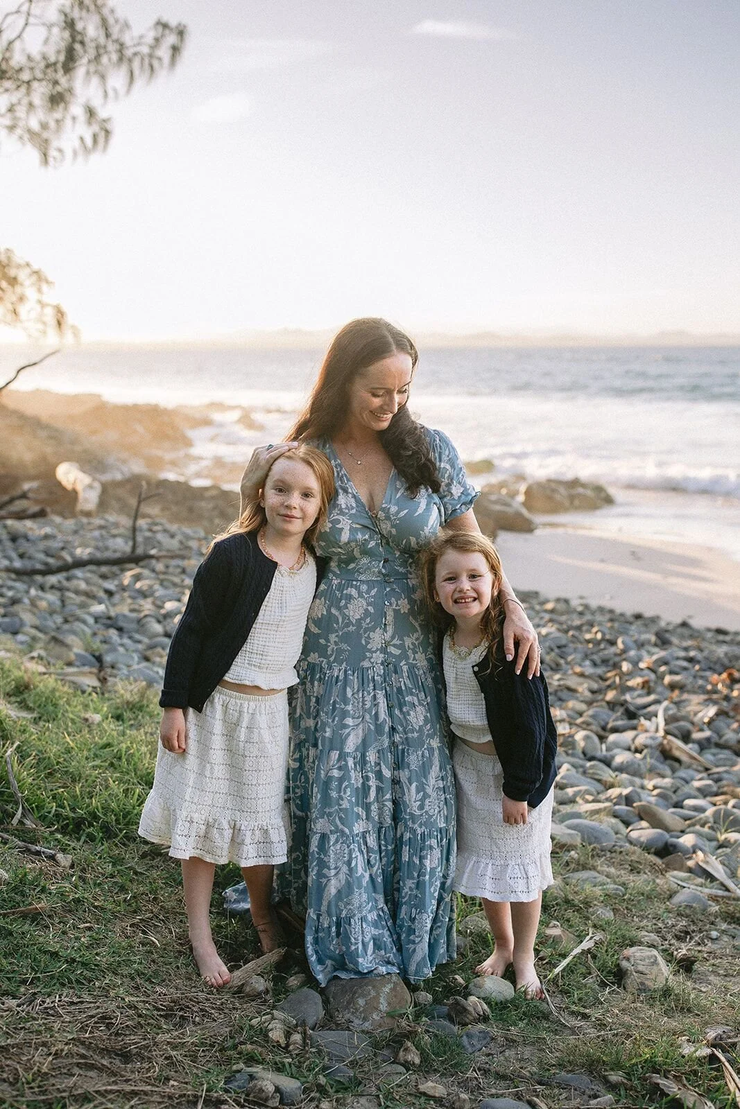 Mum holding her two daughters close and smiling on the pebbly edge of Wategos Beach