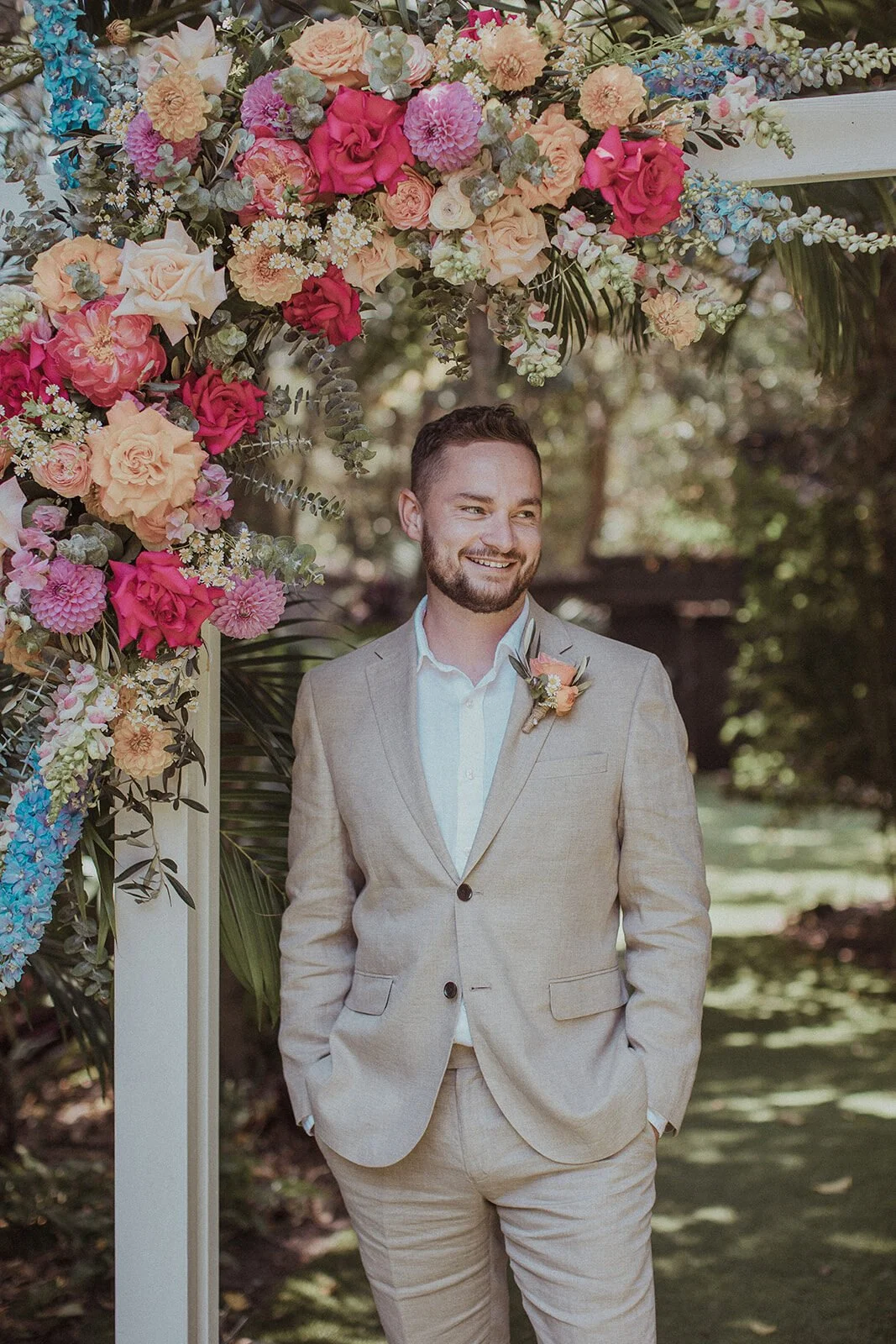 Groom in a beige suit beside a floral wedding arch, Eliza and Greg's elopement at Broken Head, Byron Bay