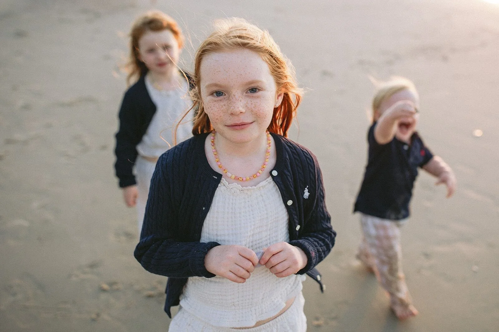 Girl with her hands clasped looking at the camera while her sister and little brother play behind her
