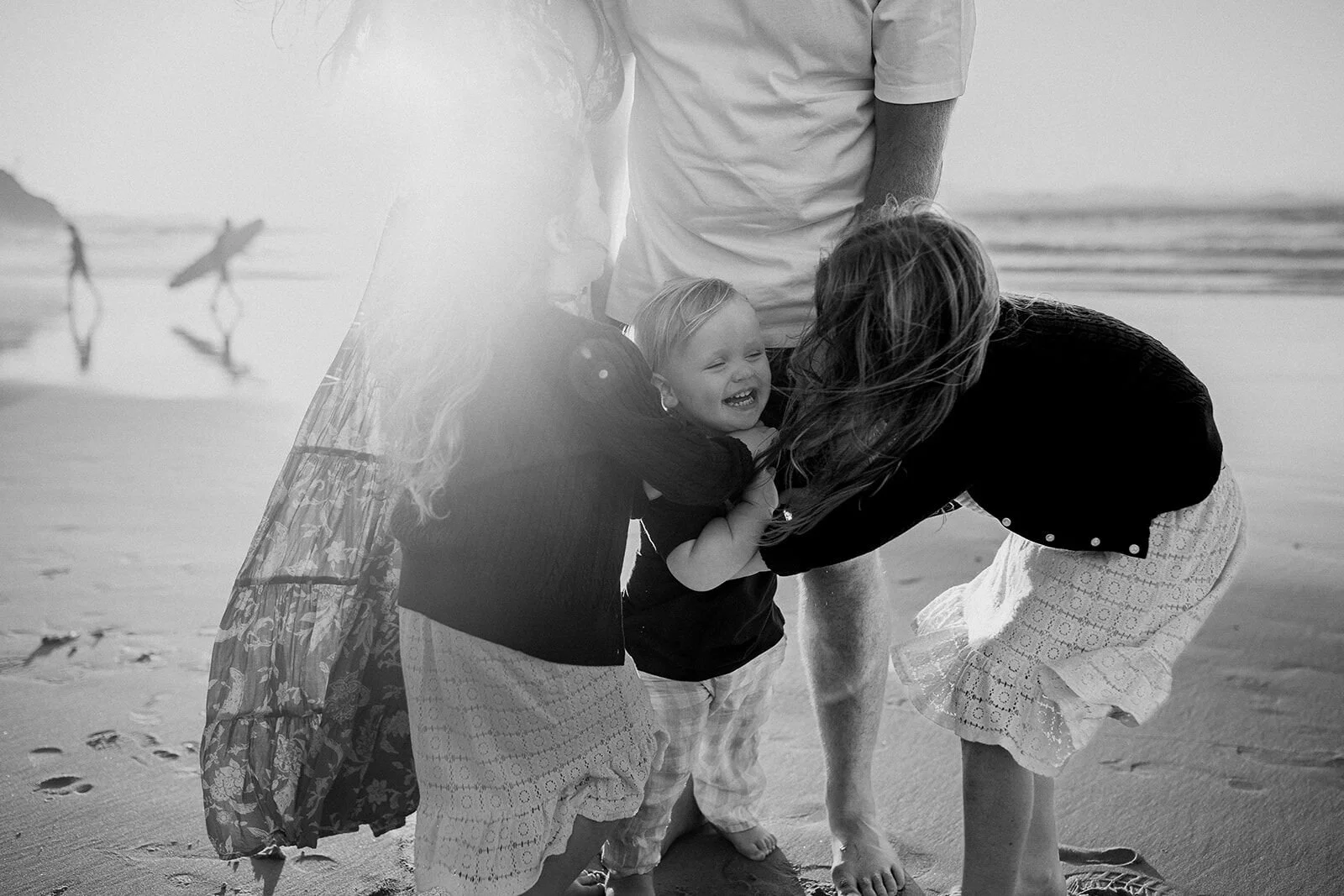 Black and white photo of a toddler and his big sister hugging in front of their parents, a surfer in the background