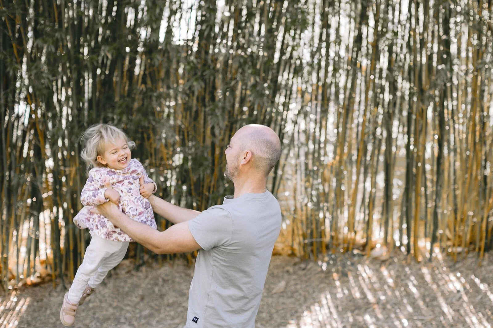 Dad swinging his laughing toddler through the air in front of bamboo, family photographer Byron Bay