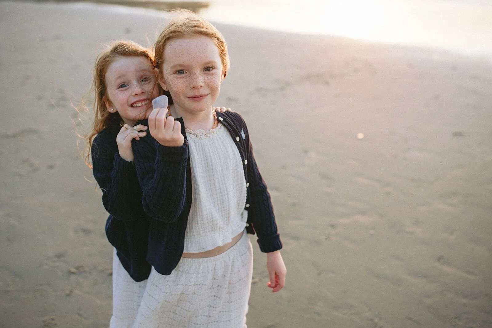 Two sisters cuddled together on the sand, one holding up a small shell