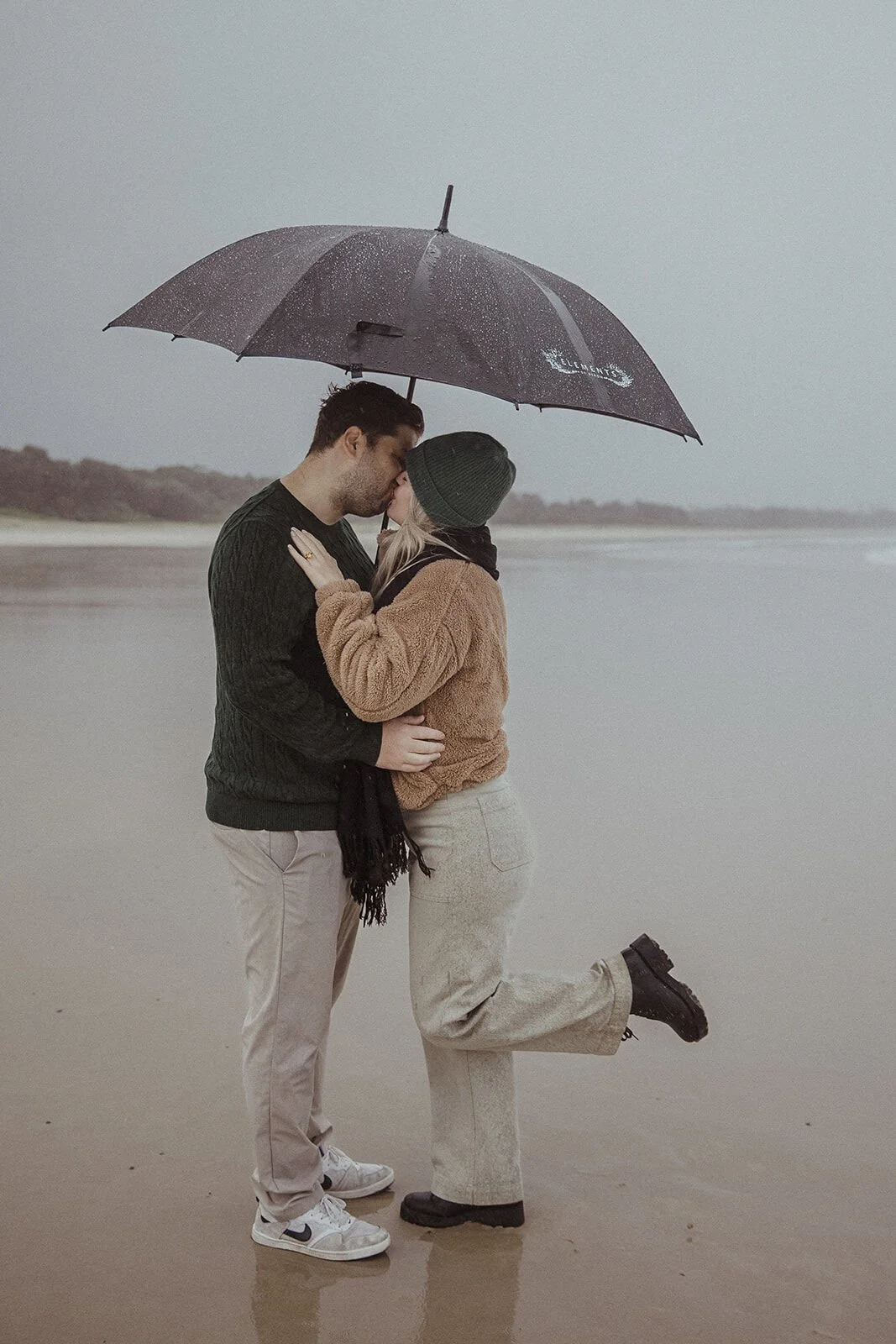 Couple kissing under the umbrella with her foot kicked up, Raphael and Vany's rainy day marriage proposal at Broken Head Beach, Byron Bay