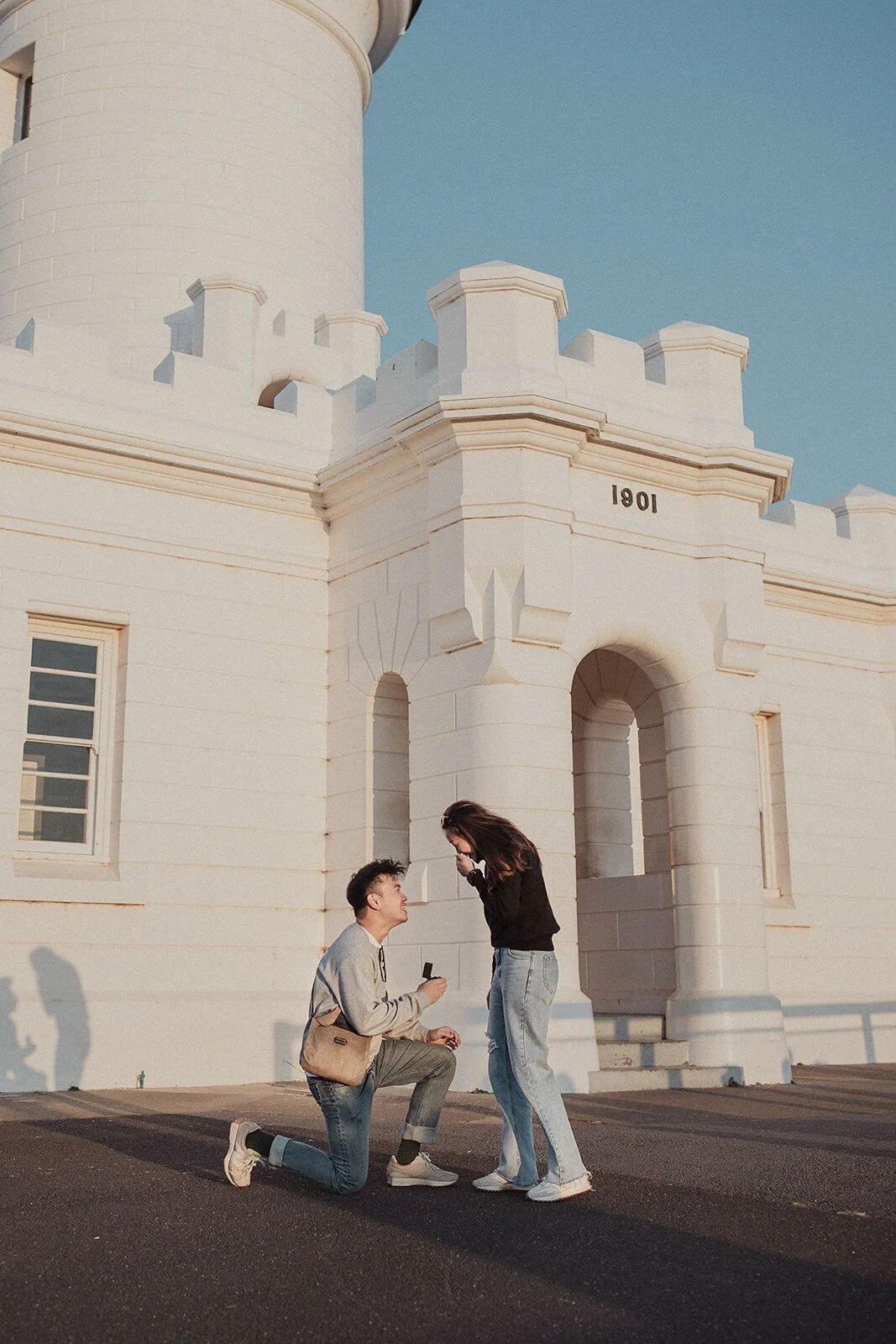 Man kneeling to propose at the lighthouse buildings as the woman stands in front of him, Douglas and Ying's sunset marriage proposal at Cape Byron Lighthouse, Byron Bay