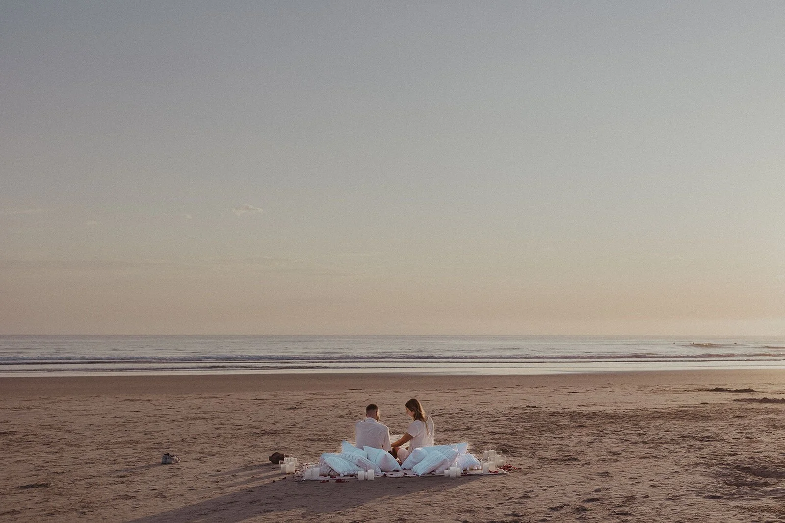 Couple tiny on the empty beach at their pillow and candle picnic, Charlie and Hannah's sunrise marriage proposal at Broken Head Beach, Byron Bay