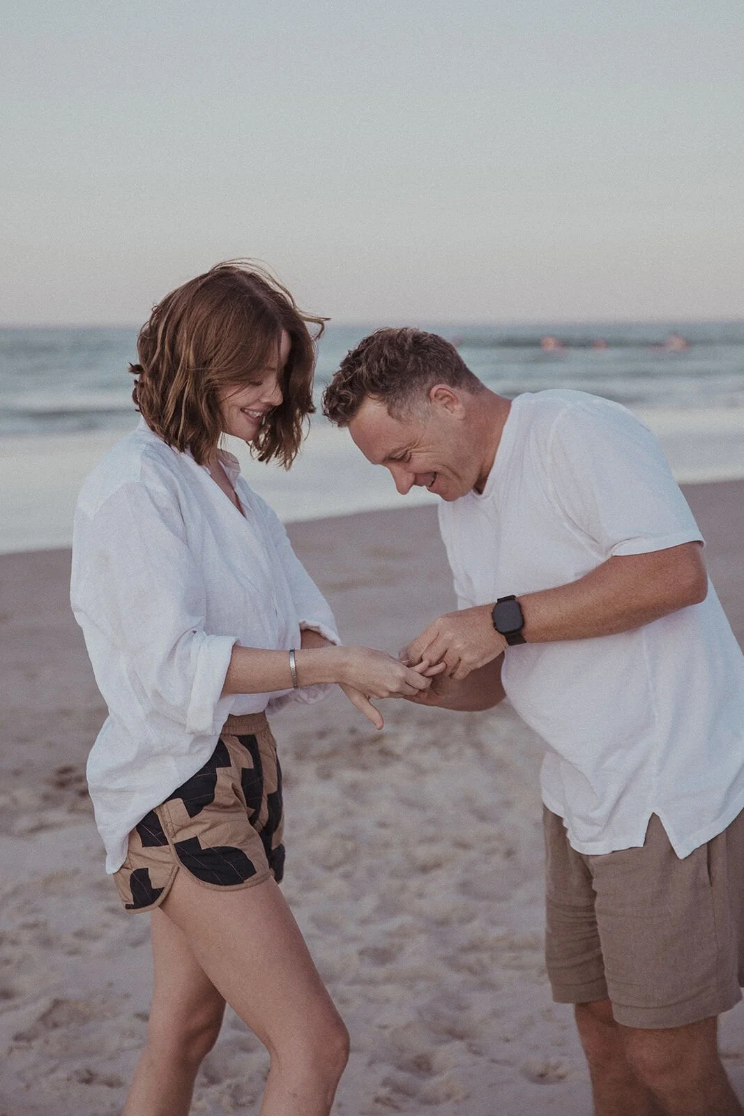 Man sliding the ring onto her finger as they smile at each other, Jamie and Jessica's beach marriage proposal at Belongil Beach, Byron Bay