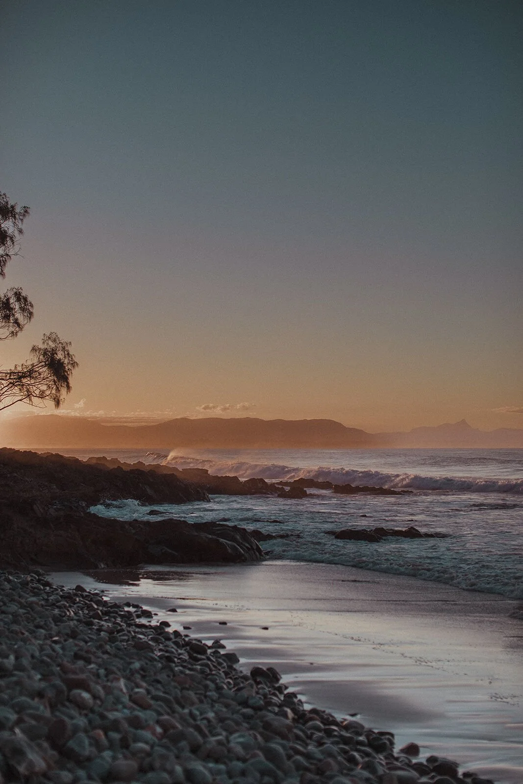 Coastline at dusk with a tree silhouette and pebble beach, John and Emma's sunset marriage proposal at Wategos Beach, Byron Bay