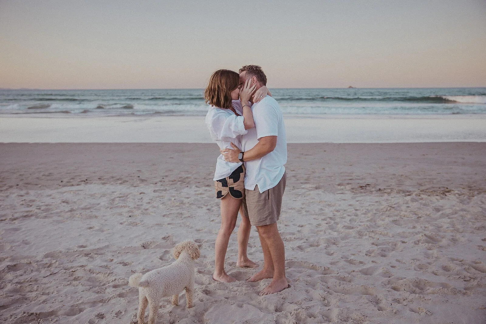 Couple kissing with their dog watching, Jamie and Jessica's beach marriage proposal at Belongil Beach, Byron Bay