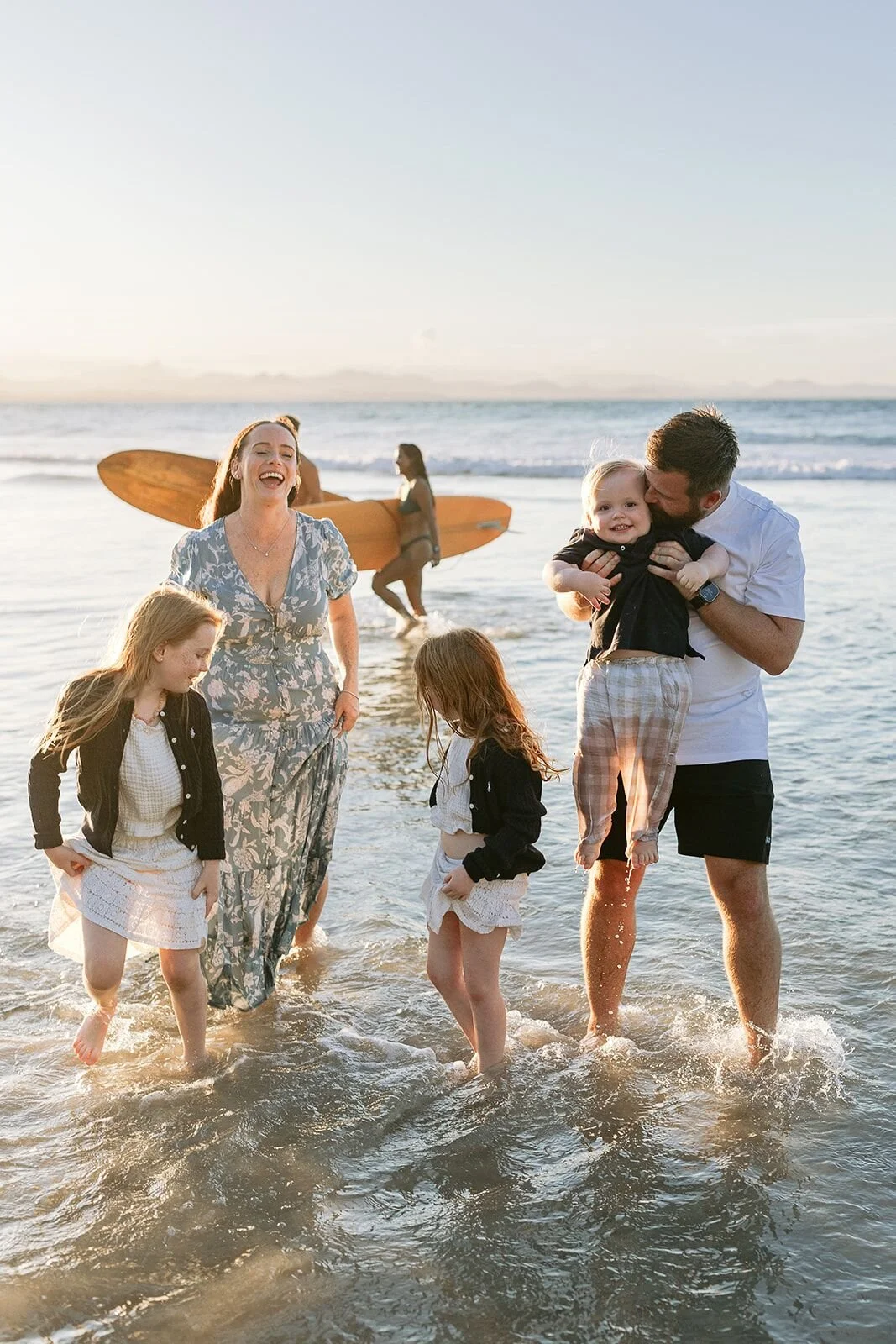 Family walking through the shallows at Wategos Beach, dad carrying the toddler, a wooden surfboard behind them