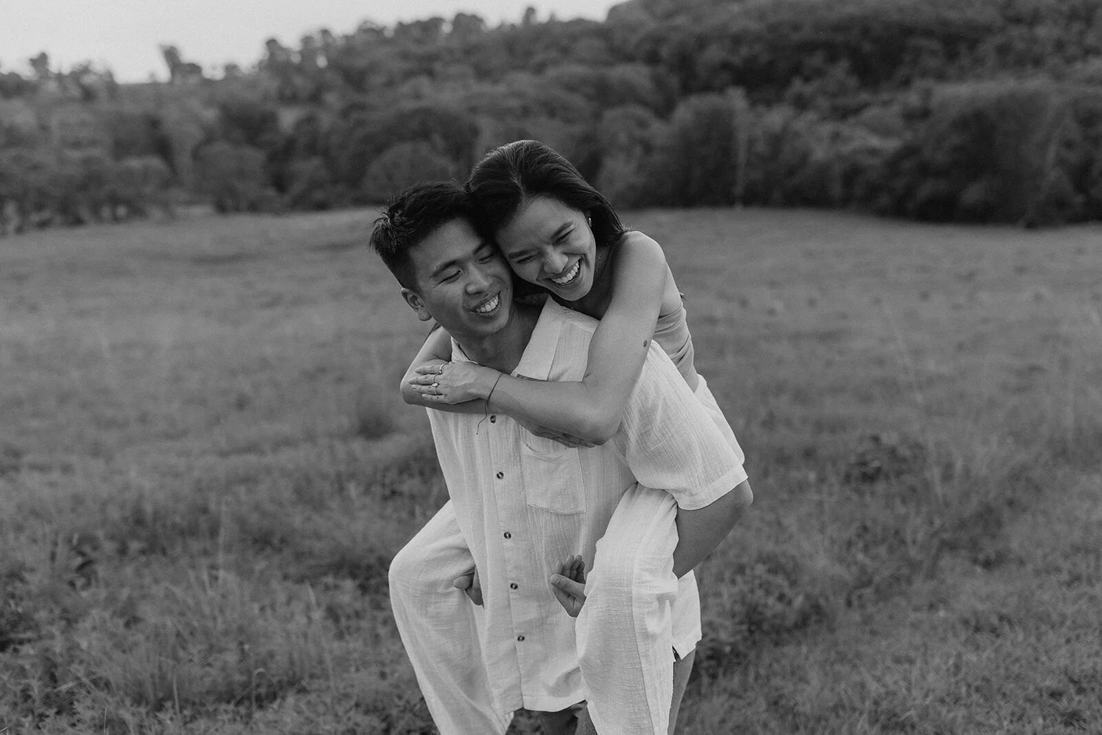 Black and white of the couple laughing during a piggyback in the paddock, Darien and Jacqui's picnic marriage proposal at Heartwood Farm, Byron Bay hinterland