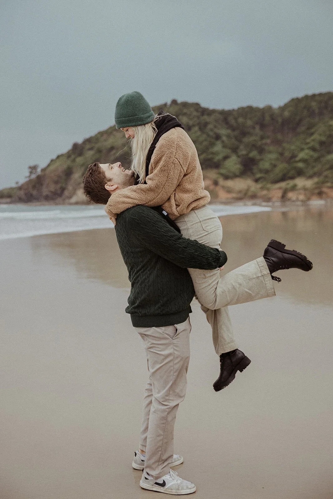 Man lifting his fiancée off the sand in the rain at the water's edge, Raphael and Vany's rainy day marriage proposal at Broken Head Beach, Byron Bay