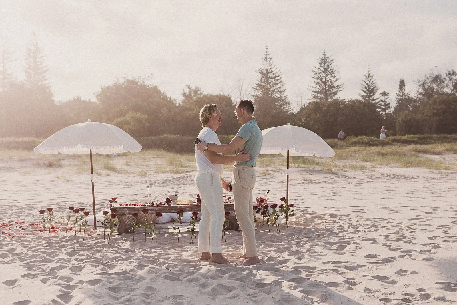 Couple holding each other on the sand with the picnic and umbrellas behind, Byron and Luke's marriage proposal at Cabarita Beach, Northern Rivers