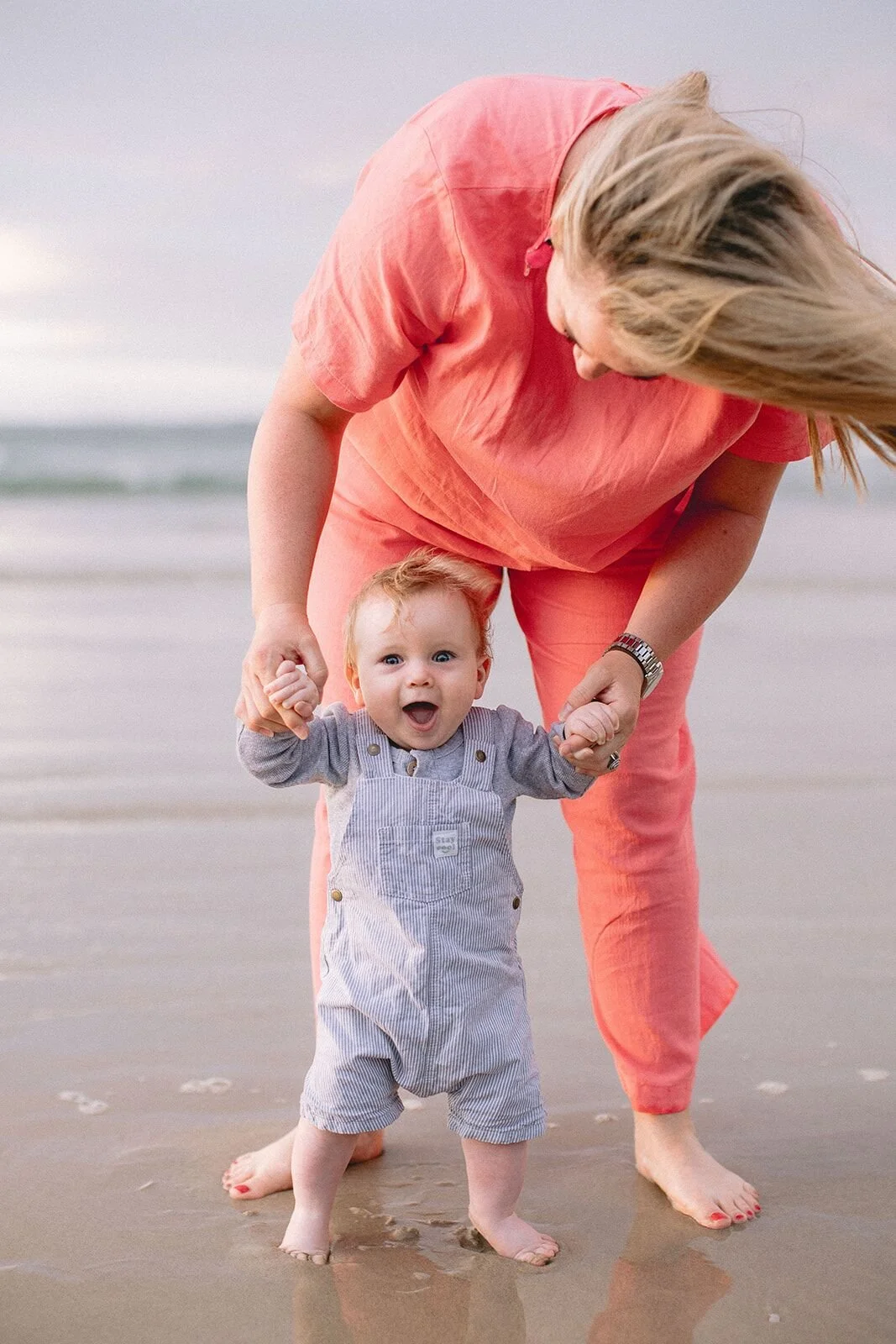 Aunt in coral guiding a baby boy taking his first steps on the sand, both laughing