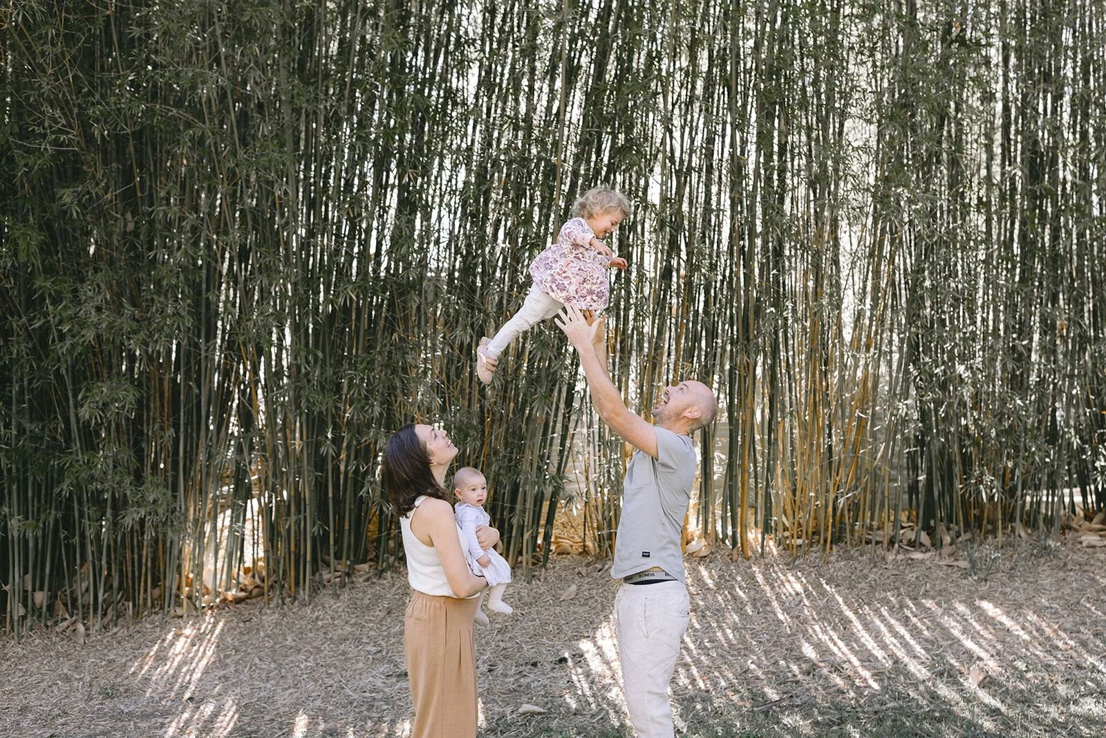 Dad lifting his toddler into the air in a bamboo grove while mum holds the baby, family photography at home near Byron Bay